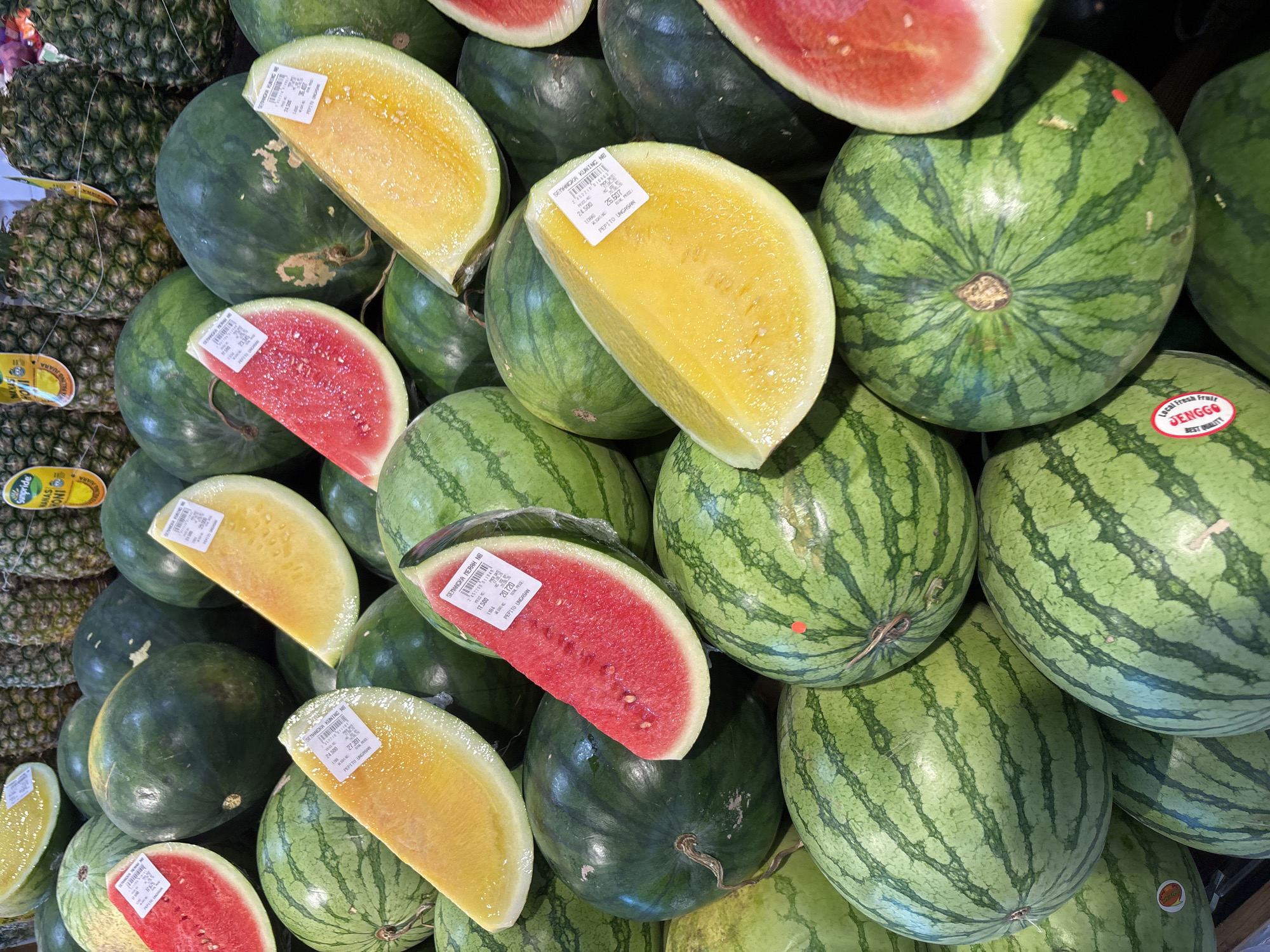 A close-up, eye-level shot features a bounty of watermelons and pineapples, likely from a market stall in Kuta Selatan, Indonesia. The watermelons, in various stages of ripeness, are piled high. Some are whole, with their characteristic dark green rind and lighter green stripes. Others are sliced into wedges, revealing their vibrant red or yellow flesh. Several wedges are individually wrapped in clear plastic, bearing white price stickers with Indonesian text. In the background, the spiky crowns of pineapples are visible, also with price tags. The scene is well-lit, highlighting the fresh produce and suggesting a bustling market environment.