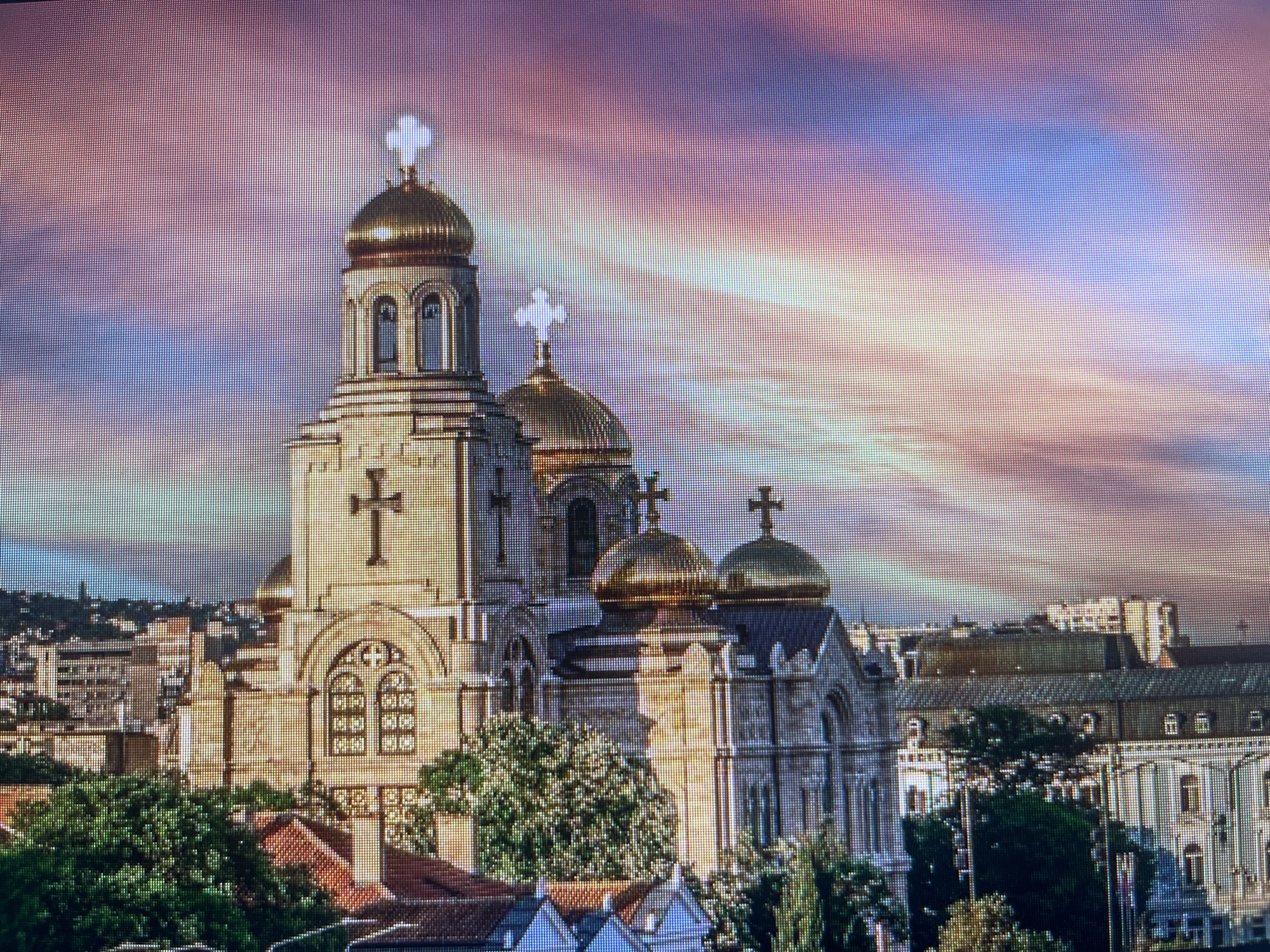 An elevated shot captures a magnificent Eastern Orthodox cathedral, likely the Dormition of the Theotokos Cathedral in Varna, Bulgaria, under a dramatically colored sky. The central subject is a large, ornate church featuring light-colored stone construction with intricate arched windows and facades. It boasts multiple golden, onion-shaped domes, each crowned with a bright white cross. Two prominent bell towers, also topped with golden domes and crosses, rise on the left side of the main structure.
The sky above is a striking blend of purple, pink, orange, and blue hues, suggesting either a vivid sunrise or sunset, with streaky clouds adding to the atmospheric effect. Below and around the cathedral, a dense urban landscape unfolds, with various buildings and abundant green trees, particularly in the foreground and flanking the church. The image offers a panoramic view of the cityscape, highlighting the cathedral as a dominant landmark. No specific activity or individuals are visible. The photograph itself displays a distinct grid pattern, indicative of being captured from a digital screen.