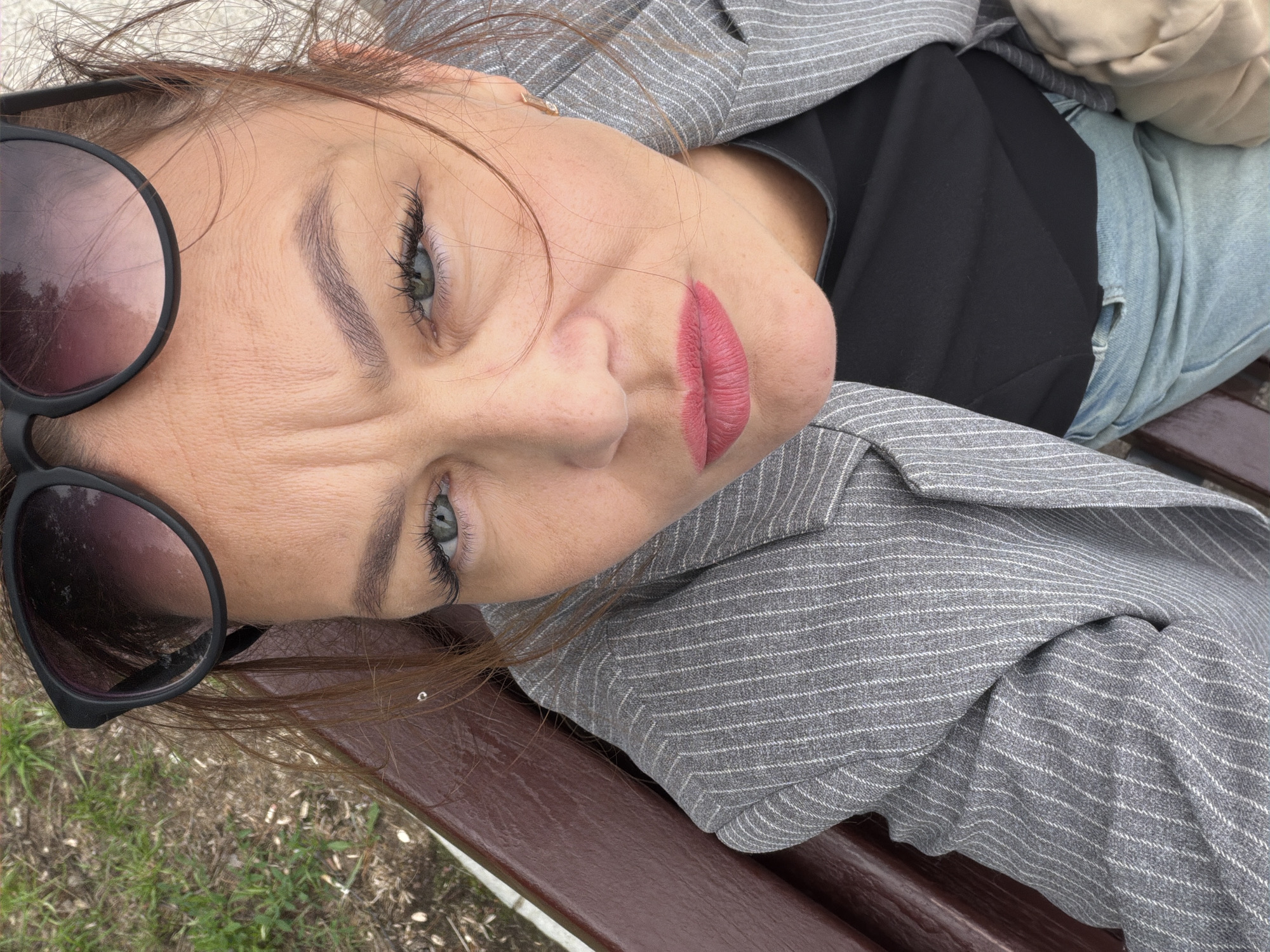 A woman wearing sunglasses, a pinstripe blazer, and a black top sits on a brown wooden park bench. Her face is close to the camera, with her gaze directed upwards. The background shows dry grass and some scattered white objects. The woman has medium-length brown hair and is wearing bright pink lipstick. The scene appears to be outdoors in a park setting, potentially in Chaikovsky, Russia, given the user's request.