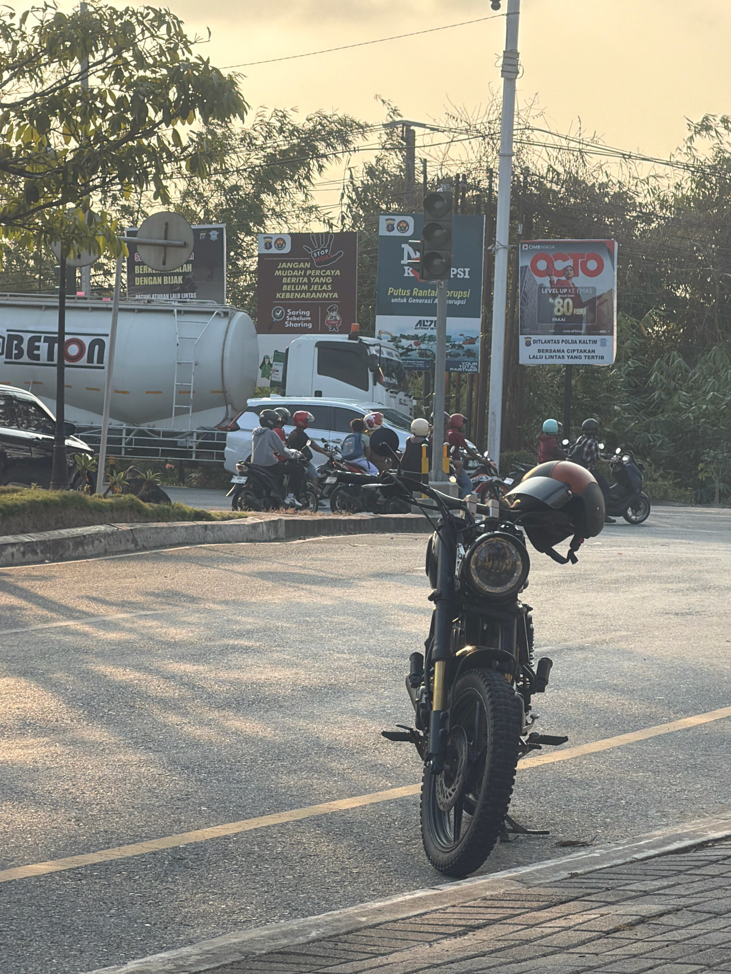 A street scene in Balikpapan, Indonesia, captures traffic and advertising. In the foreground, a black motorcycle with orange accents is parked on the side of the road, its front wheel and handlebars prominently displayed. The motorcycle has a vintage aesthetic with a round headlight and spoke wheels. Behind it, several other motorcycles and their riders are visible, some waiting at an intersection. A large white tanker truck with "BETON" written on its side is parked on the left. In the background, multiple billboards display advertisements and public service announcements. One billboard on the right promotes "OCTO," a financial service, while another on the left features a safety message from the police. The sky is bright with the soft light of either sunrise or sunset, casting long shadows across the asphalt. Lush green trees line the background, and power lines crisscross above the street. The image appears to be taken from a low angle, looking across the road towards the advertisements and traffic.