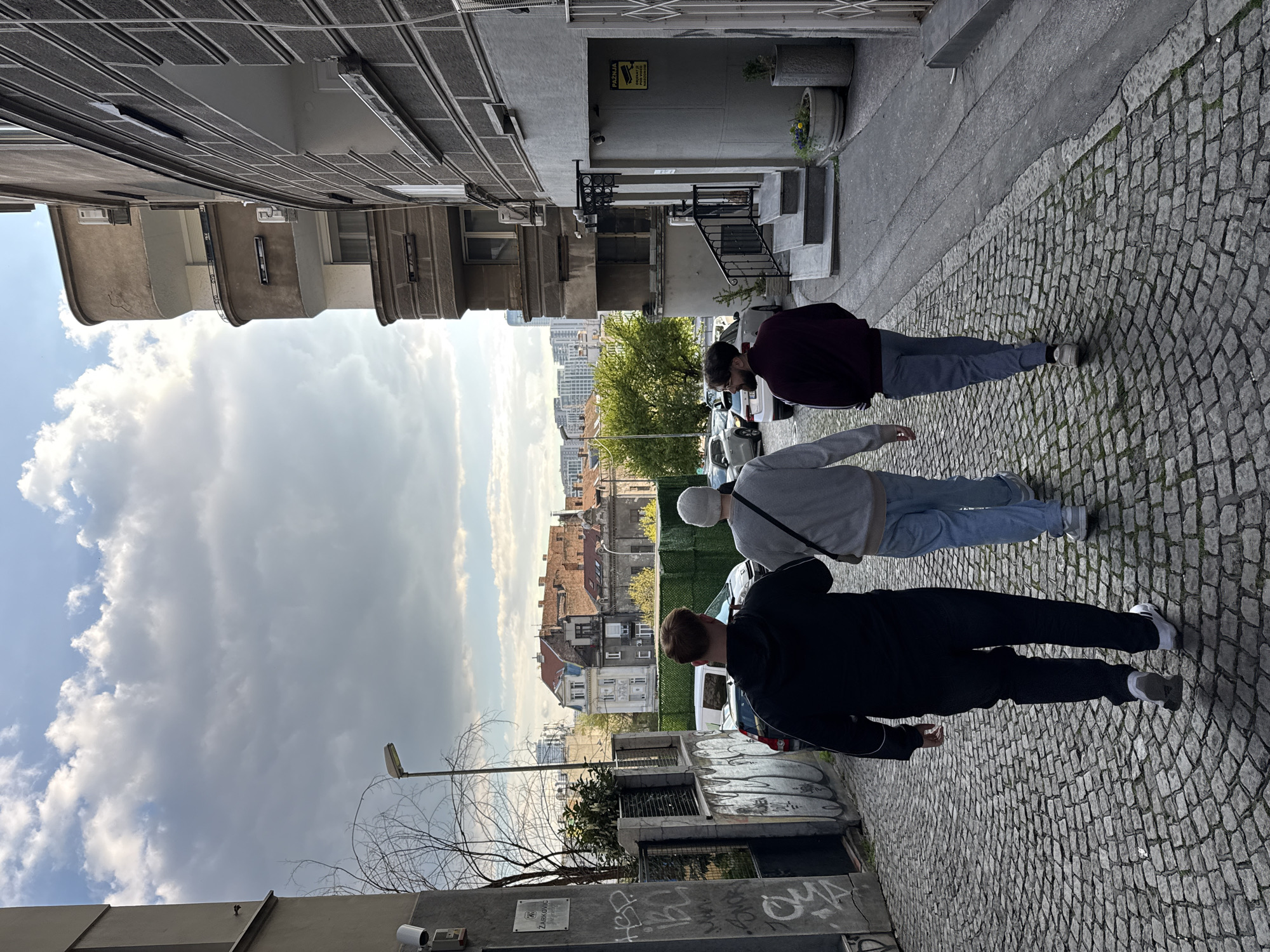 The image captures a street scene in Belgrade, Serbia. Three people are walking away from the camera along a cobblestone street lined with buildings. The buildings on the left appear older, with visible wear and tear, while the buildings on the right are more varied in style, including apartment buildings and older residences. Graffiti is visible on the lower right-hand side of the image, along with parked cars and a glimpse of greenery. In the background, a cityscape is visible under a cloudy sky. The individuals are dressed casually: one in a dark jacket and pants, another in a gray sweater and jeans, and the third in a burgundy jacket and jeans.