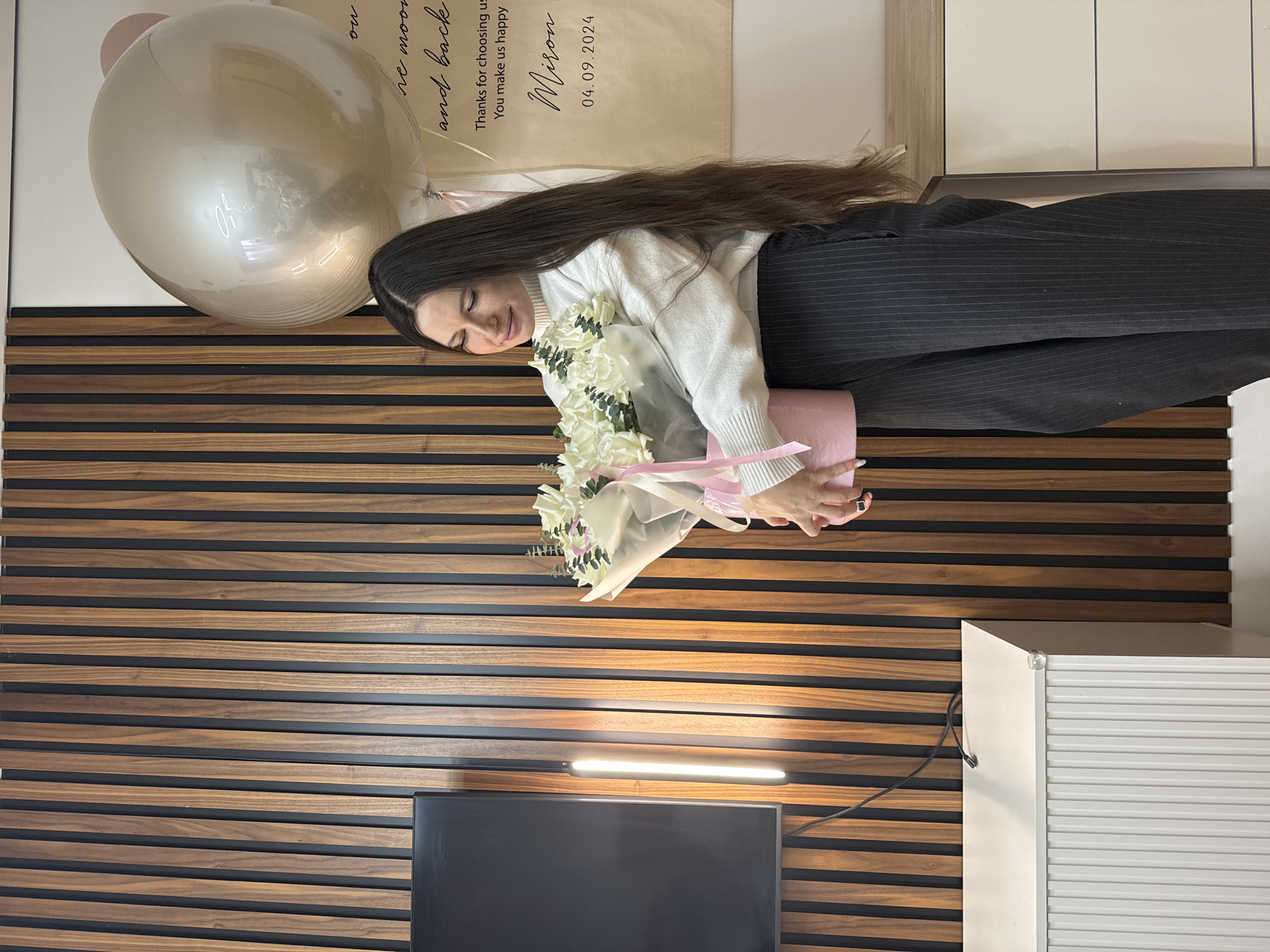 Here is a detailed description of the image:
**Scene:** The image shows a young woman holding a bouquet of white flowers. She is indoors, with a decorative wall behind her.
**People:** The main subject is a woman with long, dark hair. She is wearing a white sweater and pinstripe black pants. She appears to be smiling and looking at the flowers.
**Objects:** Key objects include:
* A bouquet of white flowers in a pink container, tied with a light pink ribbon.
* Two large balloons, one silver and one pink.
* A wall decoration with the text "Thanks for choosing us You make us happy Miron 04.09.2024".
* A brown and black striped wall.
* A black TV set.
* A picture light above the TV set.
**Actions/Interactions:** The woman is holding and looking at the bouquet of flowers. The bouquet seems to be the focal point of her attention.
**Environment:** The scene is indoors. The background wall has alternating brown and black horizontal stripes, indicating a modern interior design. There is a blank canvas with text on it, and a TV at the bottom.
**Location:** Based on the text written on the wall decoration and the data provided, it can be assumed the location is in the city of Akulovo, Gp Kubinka, Russia.