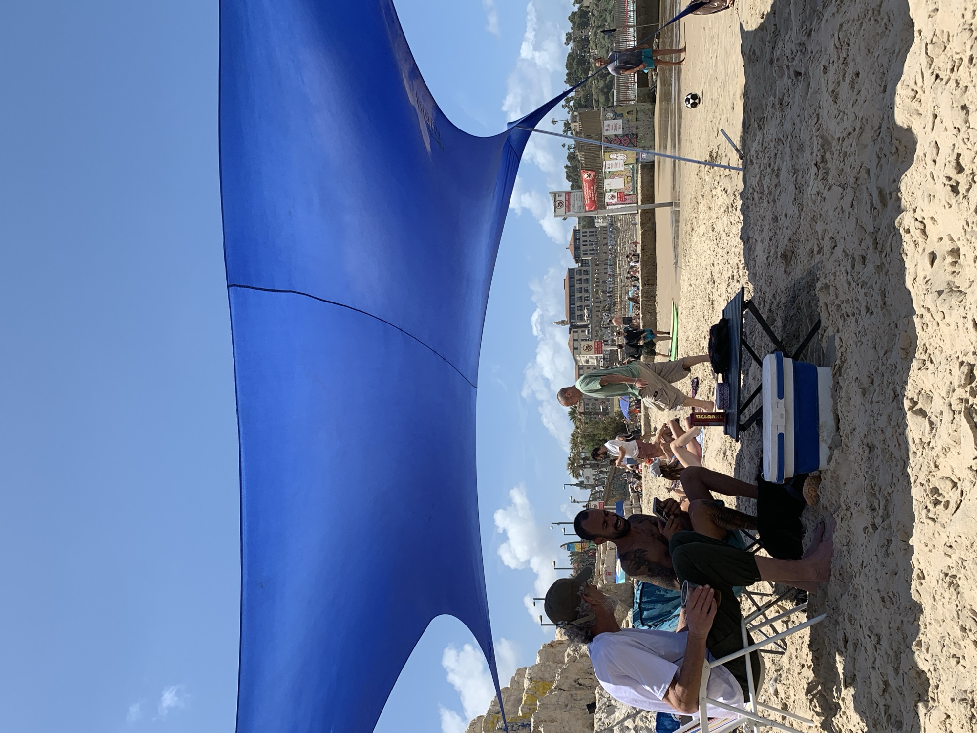 The image captures a scene on a beach in Tel Aviv, Israel, likely during a sunny day. Under a large blue sunshade, several people are seated on chairs. A man in a white shirt is sitting in the foreground. Further back, another man is looking at his cell phone. A cooler sits nearby on the sand. In the background, others are walking along the beach near a boardwalk with some buildings and palm trees in the distance. A soccer ball sits on the sand. The sand appears light in color. The sky is blue with some clouds.