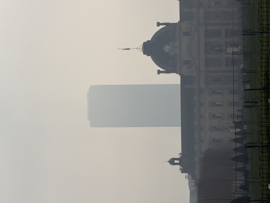 The image depicts an urban landscape in Paris, France, under conditions of dense atmospheric haze or fog. In the distant background, the Tour Montparnasse skyscraper is largely obscured, appearing as a tall, light grey rectangular silhouette with indistinct structural elements visible at its apex. In the midground, the École Militaire building extends across the lower half of the frame. This classical-style edifice features a prominent dark-domed roof on its right side, adorned with a visible clock face and topped by a flagpole displaying a dark flag. A smaller, bell-shaped cupola is present on the building's left side. The facade of the École Militaire displays multiple windows and architectural details. The foreground consists of a green grassy area containing sparse, leafless trees and conical shrubs, all partially enclosed by a series of low-lying, dark-colored temporary metal fences or barriers. The top edge of a more substantial black metal fence is visible at the very bottom of the frame. No human subjects, specific actions, or interactions are discernible.