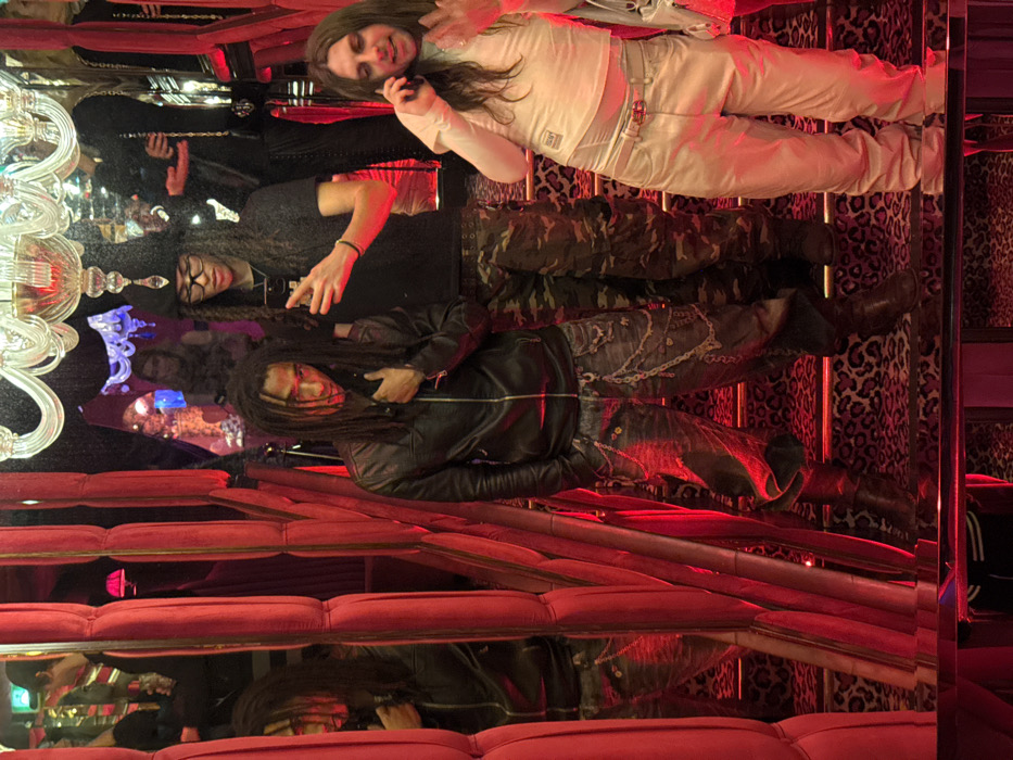 The image displays three male individuals standing on a staircase within the *Raspoutine Paris* nightclub. The environment is characterized by intense red lighting, extensive mirrored walls, and a prominent crystal chandelier overhead. The staircase features a leopard print carpet.
The individual on the left has dark dreadlocks and is wearing a black leather jacket, dark distressed jeans with multiple chains, and dark boots. Their right hand is at their chest, and they gaze downwards and to the left.
The central individual has long dark dreadlocks, wears glasses, a dark t-shirt, camouflage cargo pants, and a dark hat adorned with a white crystal-like embellishment. They hold a black smartphone in their right hand, pointed towards the viewer.
The individual on the right has long brown hair and is dressed in a white long-sleeved top, light-colored baggy pants with a belt, and carries a white bag. They are holding a white smartphone to their right ear, appearing to be engaged in a phone call, with their left hand raised in a gesture.
Reflections of the individuals and other patrons are visible in the surrounding mirrored surfaces, which are framed by red upholstered panels.