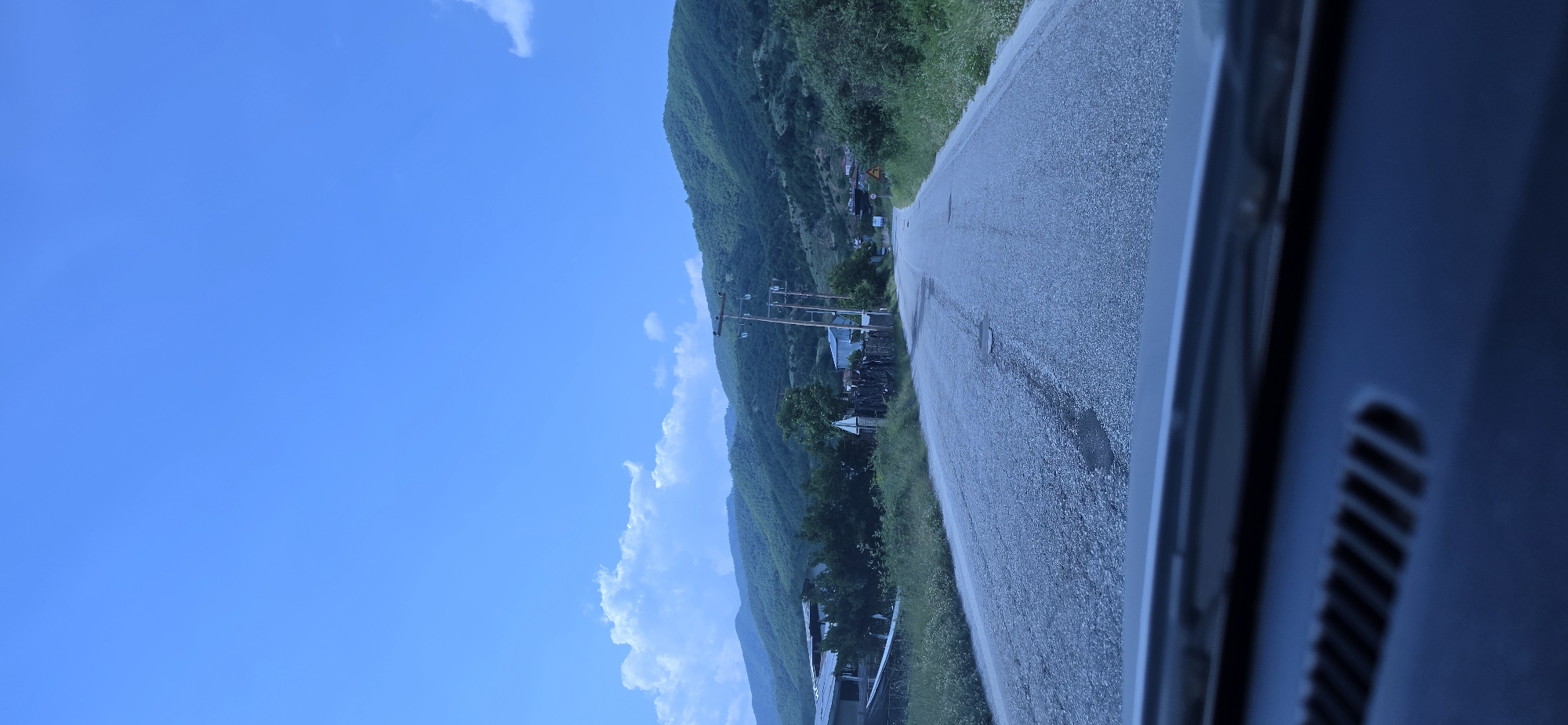 The image captures a static, daytime view of a rural road in the city of Melivoia, Greece, observed from the interior of a vehicle, with the dashboard visible at the bottom edge. A two-lane asphalt road, exhibiting areas of wear and minor potholes, extends forward and curves gently to the right in the distance. The road is bordered on both sides by grassy shoulders and dense green vegetation, including trees and bushes. A series of wooden utility poles with overhead lines runs parallel to the road on both the left and right sides. In the background, multiple layers of verdant, tree-covered hills and mountains are visible. The sky is bright blue, featuring scattered white cumulus clouds, with a more substantial cloud formation present above the left mountainous area. A yellow diamond-shaped road sign, its specific symbol unclear due to distance, is situated on the right side of the road further down. No people or active subjects are discernible in the scene.