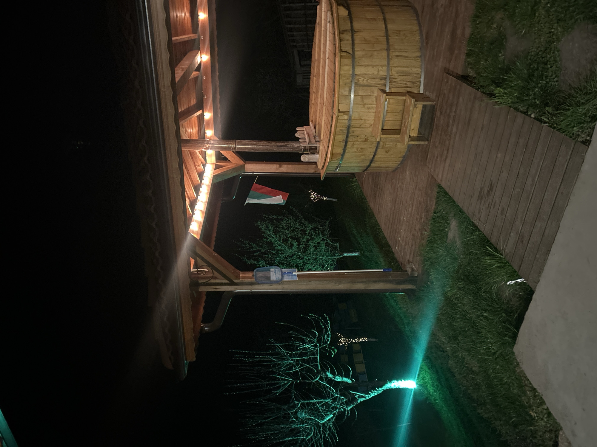The media file displays an outdoor scene at night, likely in Zlatograd, Bulgaria, illuminated by various artificial light sources. The primary subjects include a large, circular wooden hot tub or plunge pool with metal bands, featuring a small set of wooden steps. Adjacent to it is a rustic wooden pergola or shelter, constructed with exposed beams and adorned with warm-toned string lights, casting a glow on the structure.
A wooden plank pathway extends from the hot tub area across a grassy patch. A small Bulgarian flag (white, green, and red stripes) hangs from a pole near the wooden structure, serving as a clear location cue. A clear plastic bottle or container is visible hanging from one of the pergola's posts.
The foreground and midground feature a bare, leafless tree, dramatically lit by a strong green light source emanating from the ground, creating a vibrant teal glow that contrasts with the warm string lights. More grass and obscured vegetation are visible around the pathway and in the distance. The overall setting suggests a tranquil, inviting space for evening relaxation or leisure, possibly at a lodge or guesthouse. There are no people visible, and no discernible text.