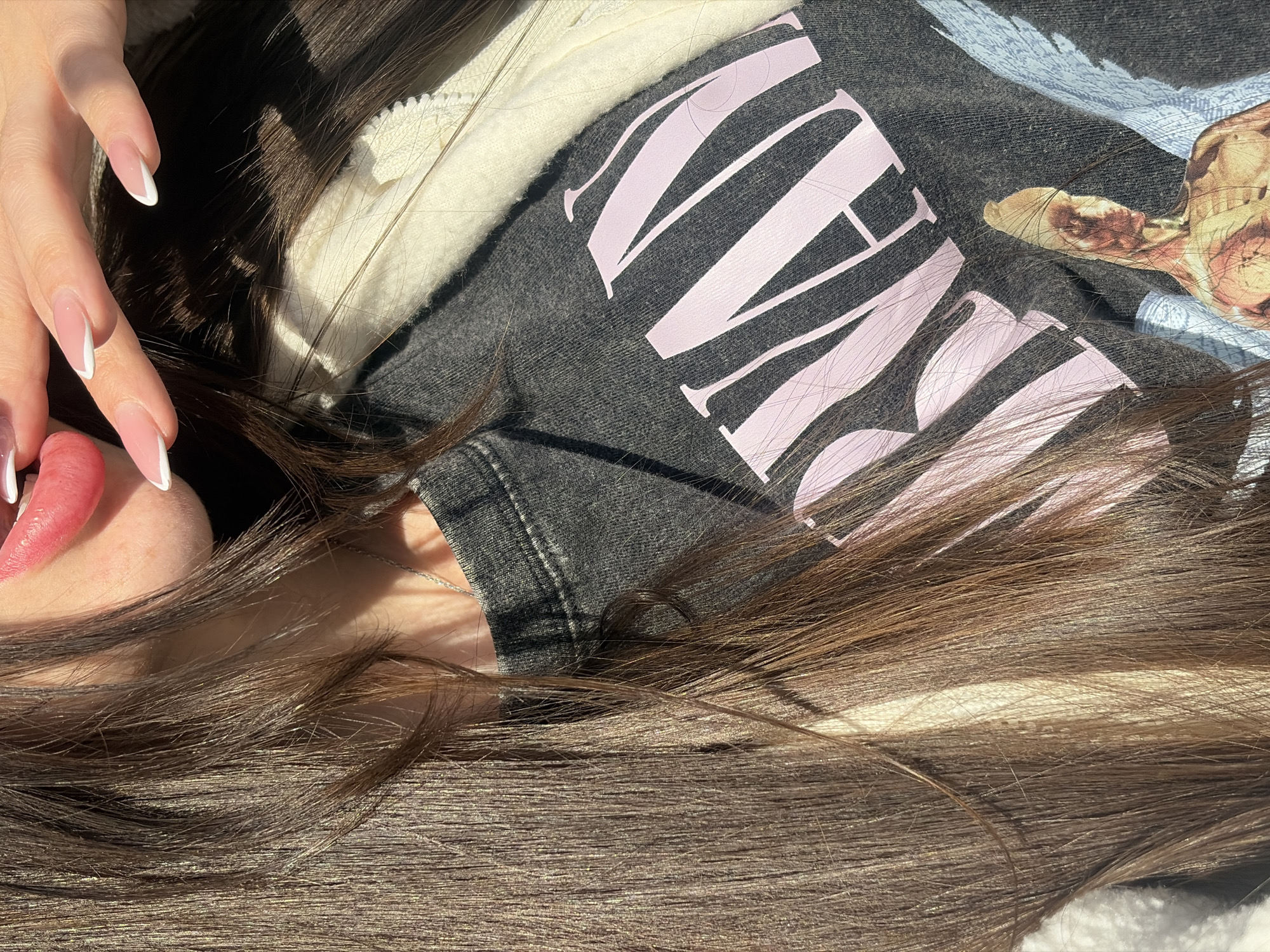 A close-up, mid-torso portrait captures an individual under direct sunlight. The subject, visible from the lower lip to the upper chest, has long, straight, dark brown hair with lighter brown highlights, draped over the left shoulder and chest. The lower lip is painted with bright pink lipstick and is slightly parted, revealing a sliver of white teeth. A thin silver chain necklace is visible around the neck.
The individual's right hand, with nails featuring a French manicure (pink base, white tips), is positioned with fingers touching the lower lip and chin area. The subject wears a dark gray, faded t-shirt featuring large, light pink letters spelling "NIRVAN" (part of "NIRVANA") on the chest. Below this text, a graphic depicts a yellow-toned, anatomically rendered male figure with prominent musculature and light blue wings, consistent with the "In Utero" album artwork. Over the t-shirt, a cream or off-white fuzzy jacket, possibly fleece, with a visible zipper, is worn. Strong sunlight casts distinct shadows across the neck and clothing.
The specific location within the city of Saint Petersburg, Russia, is not identifiable from the visual content of this image.