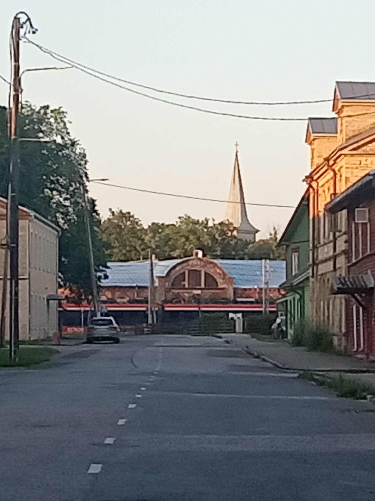 The image shows a street scene in Tapa, Estonia. A paved road runs through the center of the frame, with white dashed lines marking the lanes. On the left side of the road, a multi-story building with a light-colored facade and evenly spaced windows is visible, with a tall pole topped with what appears to be a street light or utility fixture to its left. Across the street, a long, low building with a distinctive arched roof section is present, featuring red and dark horizontal banding and what appears to be large windows or display areas. Further down the street on the right, several older, multi-story buildings with pitched roofs in various colors, including green and red, line the sidewalk. A slender church spire with a cross at its apex rises above the buildings in the background. The sky is a pale, hazy blue, suggesting either early morning or late evening light. A silver car is parked on the left side of the road, facing away from the camera, and a person on a bicycle is visible on the right sidewalk. Trees with lush green foliage are interspersed throughout the scene, framing the buildings and contributing to the overall urban landscape.
