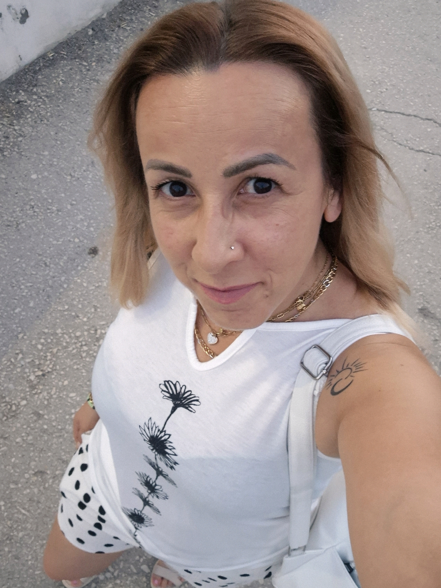 In an outdoor, overhead shot, a woman stands on a cracked, grey asphalt surface, possibly a sidewalk or street. She has fair skin, shoulder-length blonde hair, and is looking towards the camera. Her left eye is open, and her right eye is slightly closed. She has a nose ring and is wearing a white sleeveless top with a black floral design. Around her neck, she has a layered gold necklace with a heart-shaped pendant. She has a tattoo of a sun and crescent moon on her left shoulder. Her lower body is adorned with a white skirt or shorts featuring black polka dots. A white bag is slung over her left shoulder, and she is wearing light-colored sandals. The background consists of the grey asphalt, suggesting an urban environment.