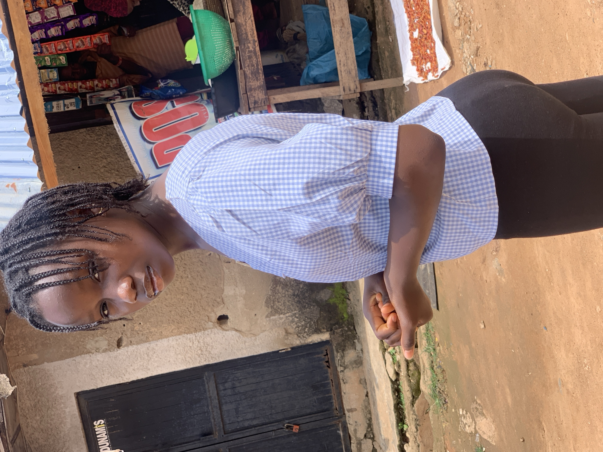 In Sabon Tasha, Nigeria, a young Black female stands outdoors in front of a building and a market stall. She has dark skin and her hair is styled in braids. She is wearing a light blue and white checkered short-sleeved shirt and black leggings. Her left hand is clasped in front of her, and her right hand is partially visible. Behind her, a dark wooden door with a red lock is set into a concrete wall. To her right, a market stall displays various goods, including packaged snacks hanging from the roof and a green basket filled with items. A wooden table holds more goods, and blue tarps are visible underneath. In the foreground, a white sheet spread on the ground is covered with dried red items, possibly chilies or small fish. The ground is covered in reddish-brown dirt and some scattered green vegetation.