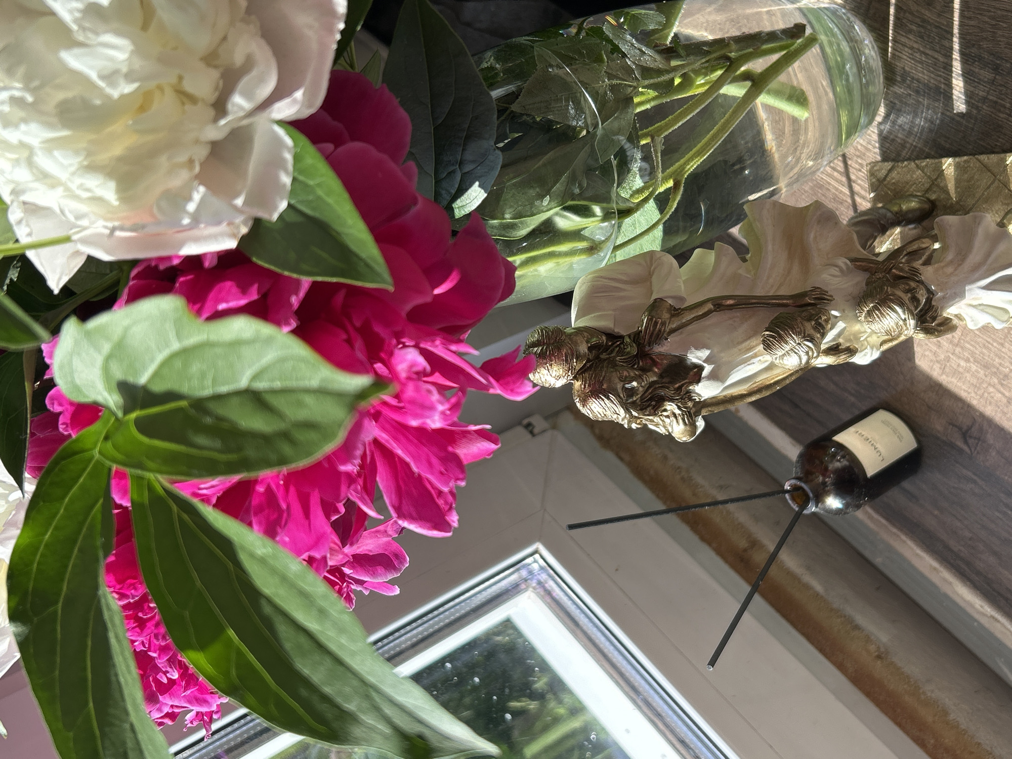 A still life photograph captures a close-up view of a bouquet of peonies, featuring both white and bright pink blooms, interspersed with lush green leaves. The flowers are arranged in a clear glass vase, visible behind the foliage. To the right of the vase, a small decorative item with golden figures, possibly a sculpture or ornament, rests on a wooden surface. A brown glass bottle with a label that reads "LUMIERE" is placed next to the ornament, with two black diffuser sticks inserted into its opening. The scene is set on a wooden surface, likely a windowsill, as a white window frame is visible in the lower part of the image. Sunlight streams in, casting distinct shadows across the wooden surface, suggesting it is daytime. The overall mood is serene and natural, highlighting the beauty of the flowers and decorative elements. The location is Chaikovsky, Russia.