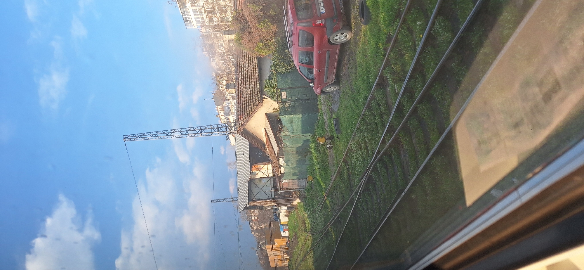 View from a train window on a bright, sunny day. A red car is parked near train tracks overgrown with vegetation. A building, possibly residential or industrial, stands adjacent to the tracks, with a construction crane visible in the background against a blue sky with scattered clouds. Urban buildings are visible in the distance. The reflection of the scene is apparent on the train window, and it's likely in or near Burgas, Bulgaria.