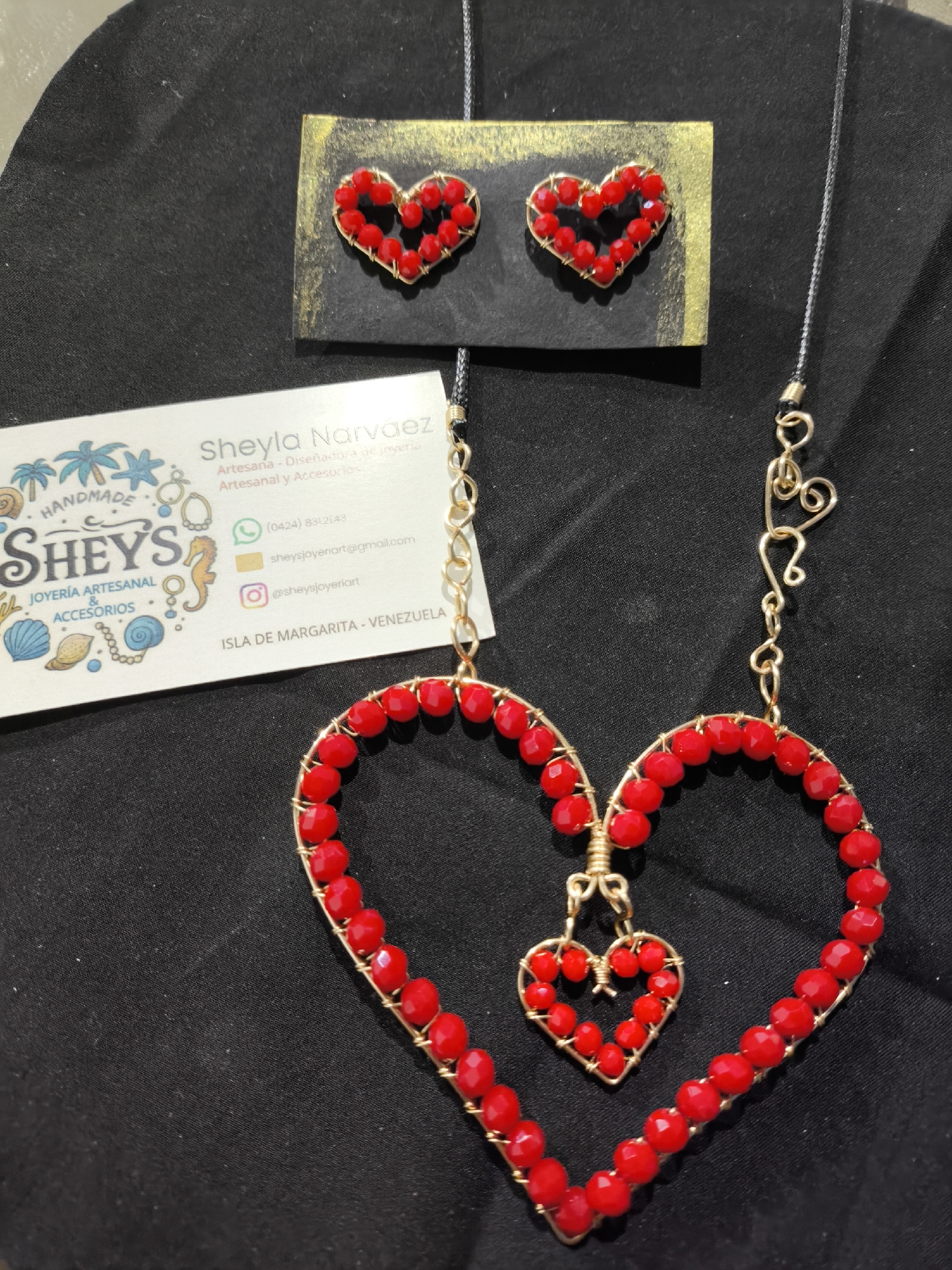 A close-up shot shows handmade jewelry displayed on a black fabric surface. The jewelry consists of a pair of heart-shaped earrings with red beads and gold wiring, and a necklace with a large heart pendant outlined with the same red beads and gold wiring, inside of which is a smaller heart pendant matching the earrings. Next to the jewelry, there is a business card for "Sheyla Narvaez," identifying her as an artisan and jewelry designer. The card includes a logo featuring a seahorse, seashells, and palm trees, along with contact information like a phone number, email address, and Instagram handle (@sheysjoyeriart). The card also notes the location: "Isla de Margarita - Venezuela." There is no indication of the city of Los Chacos in the image.