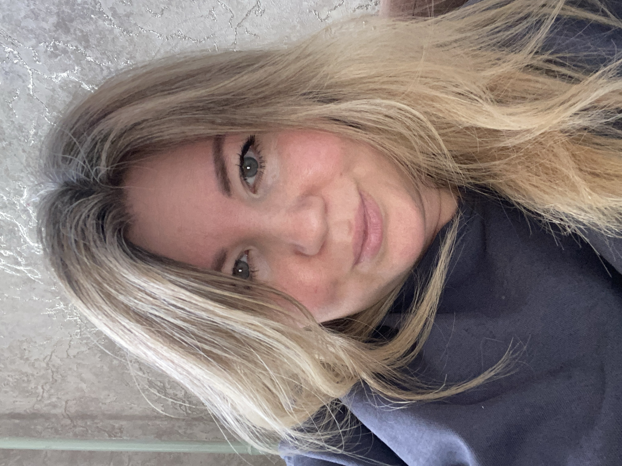 This image is a medium close-up portrait of a woman, presented with a 90-degree clockwise rotation. She appears to be an adult with fair skin and light-colored eyes, likely green or hazel, looking directly at the camera with a soft, pleasant smile. Her long, blonde hair, featuring both lighter strands and darker roots or lowlights, cascades around her face and shoulders. She is wearing a casual, dark blue or grey hooded top or sweatshirt. The background consists of an indoor wall with a textured, light grey and silver shimmering pattern, suggesting decorative wallpaper or paint. The lighting appears to be ambient indoor light. The setting is indoors, in Chaikovsky, Russia. There is no visible text in the image.
