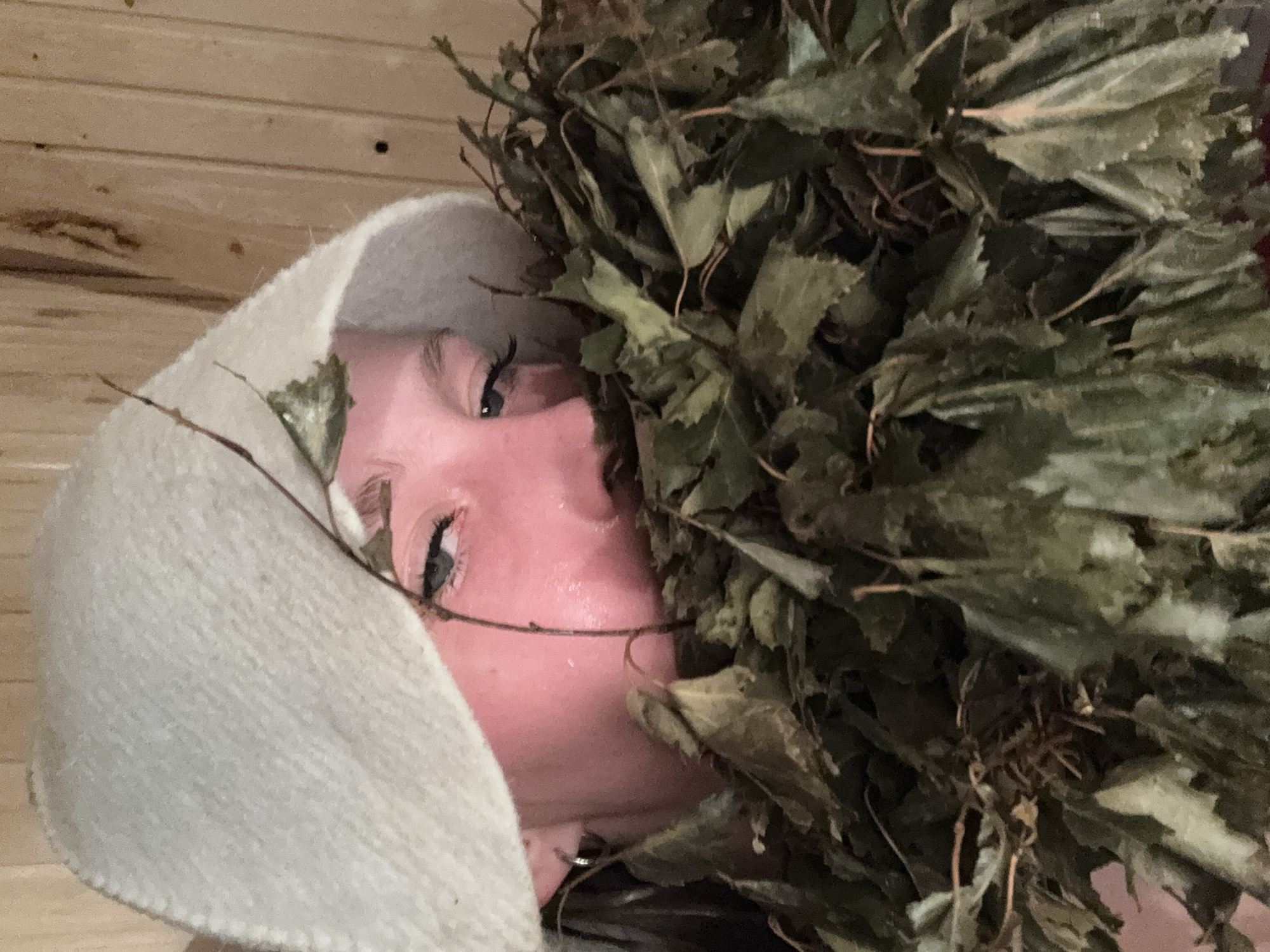 A close-up, rotated view captures a woman partially obscured by a bundle of dried branches and leaves. The woman's face is flushed, and she is wearing a light-colored, hooded head covering. Her eyes are visible, and she appears to be looking towards the camera. The background consists of wooden panels. There is no information in the picture to suggest it was taken in the city of Markovo, Russia.