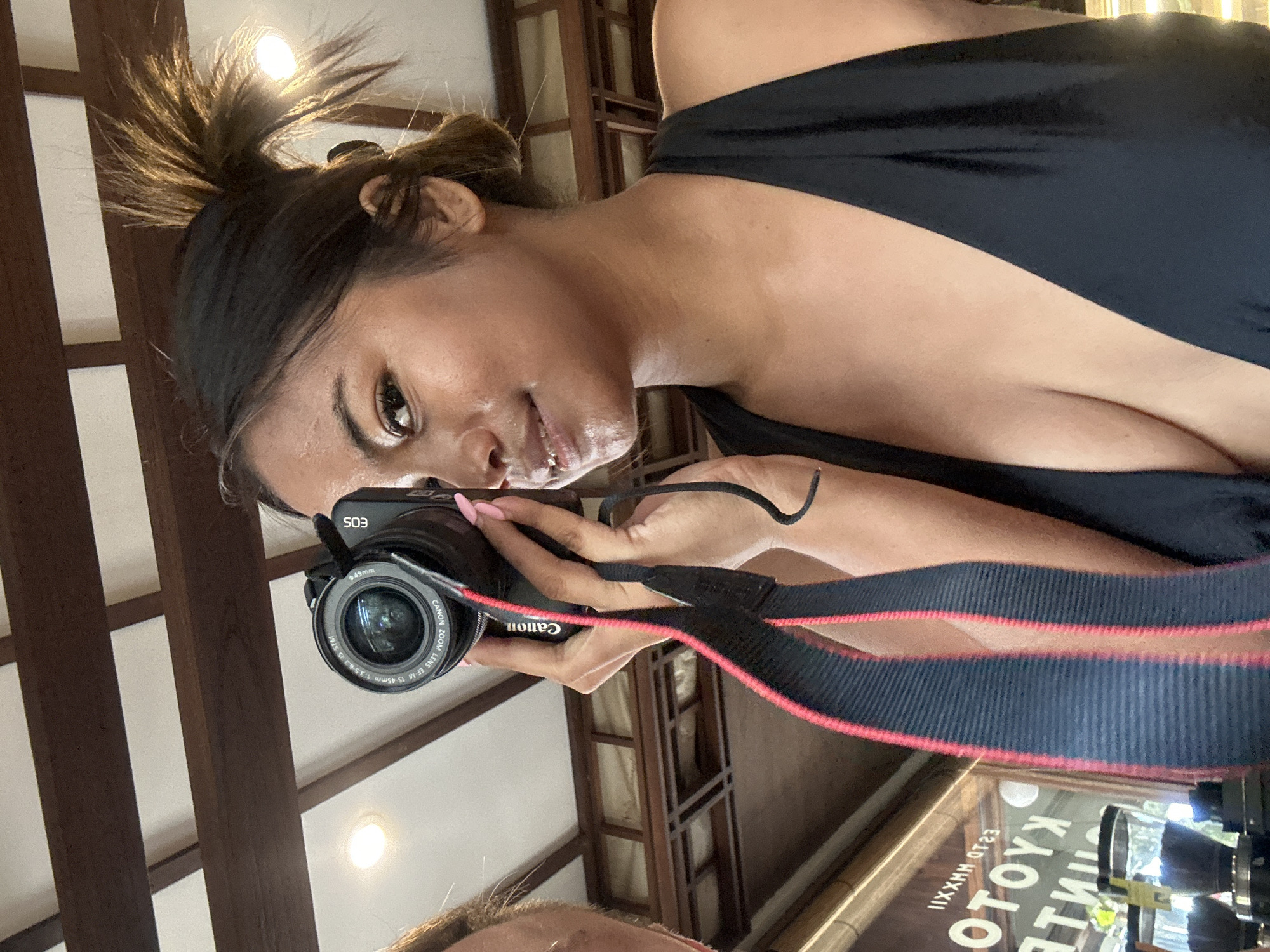 A young woman with dark hair pulled into a messy bun is taking a selfie with a Canon EOS camera. She is wearing a black halter top that reveals her cleavage. She is holding the camera with her right hand and her left hand is blurred in the foreground. The background appears to be indoors, with wooden beams on the ceiling and some bright lights. There are reflections in what appears to be a glass display case in the lower left corner, showing the words "KYOTO COFFEE" and "EST. MMXXII". This suggests the location is a coffee shop in Denpasar, Indonesia. The woman is looking at the camera with a slight smile.