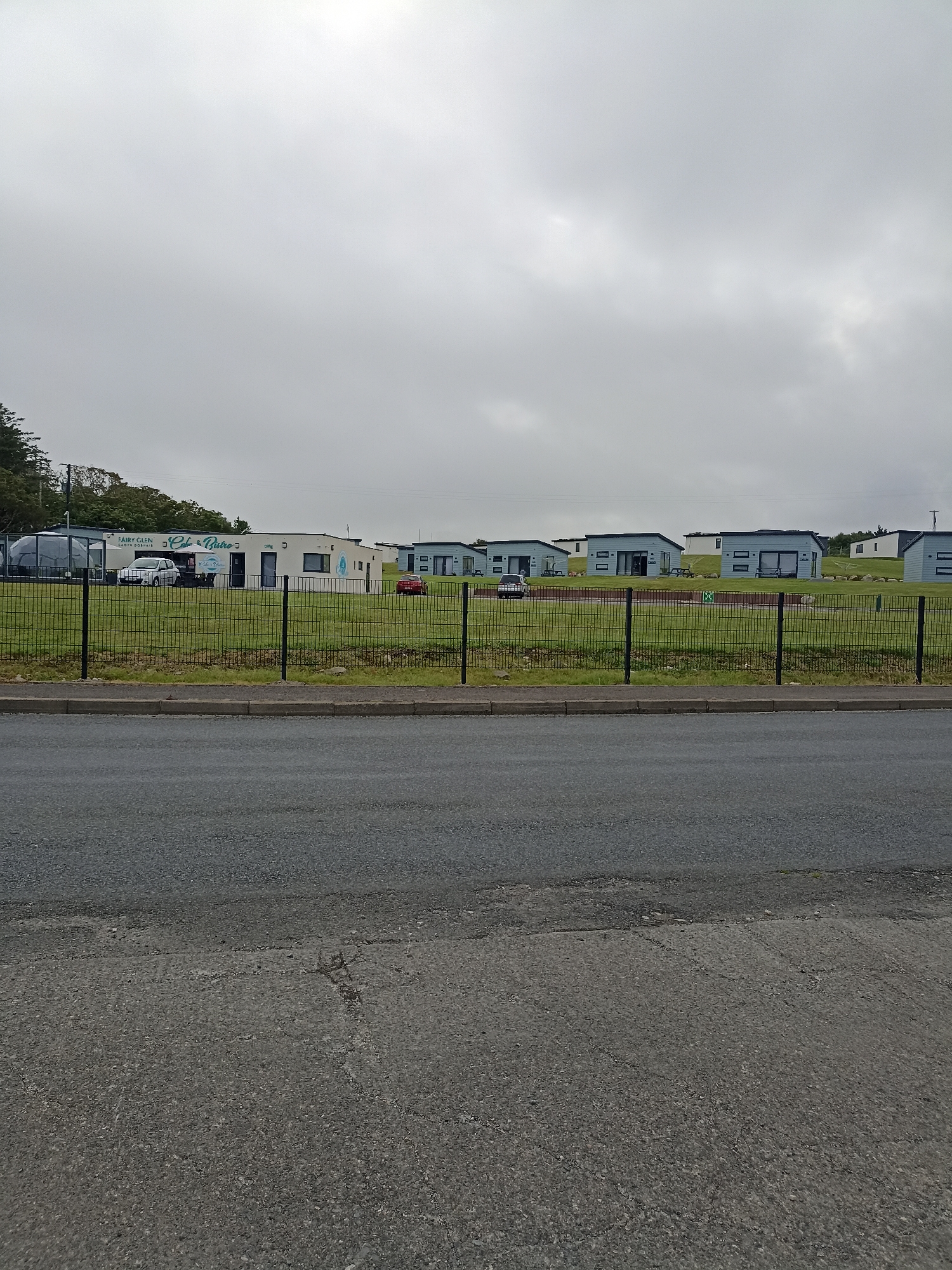 The image shows a row of modern, light-blue holiday homes with dark roofs, situated on a grassy hillside under a cloudy sky. In the foreground, a black metal fence separates a paved road from the property. To the left, there's a building with "FAIRY GLEN CAFE & BISTRO" signage and a white car parked outside. A large geodesic dome structure is also visible to the left of the cafe. The environment appears to be a holiday park or resort in Doirí Beaga, Ireland, characterized by its residential structures, a recreational building, and surrounding natural landscape.