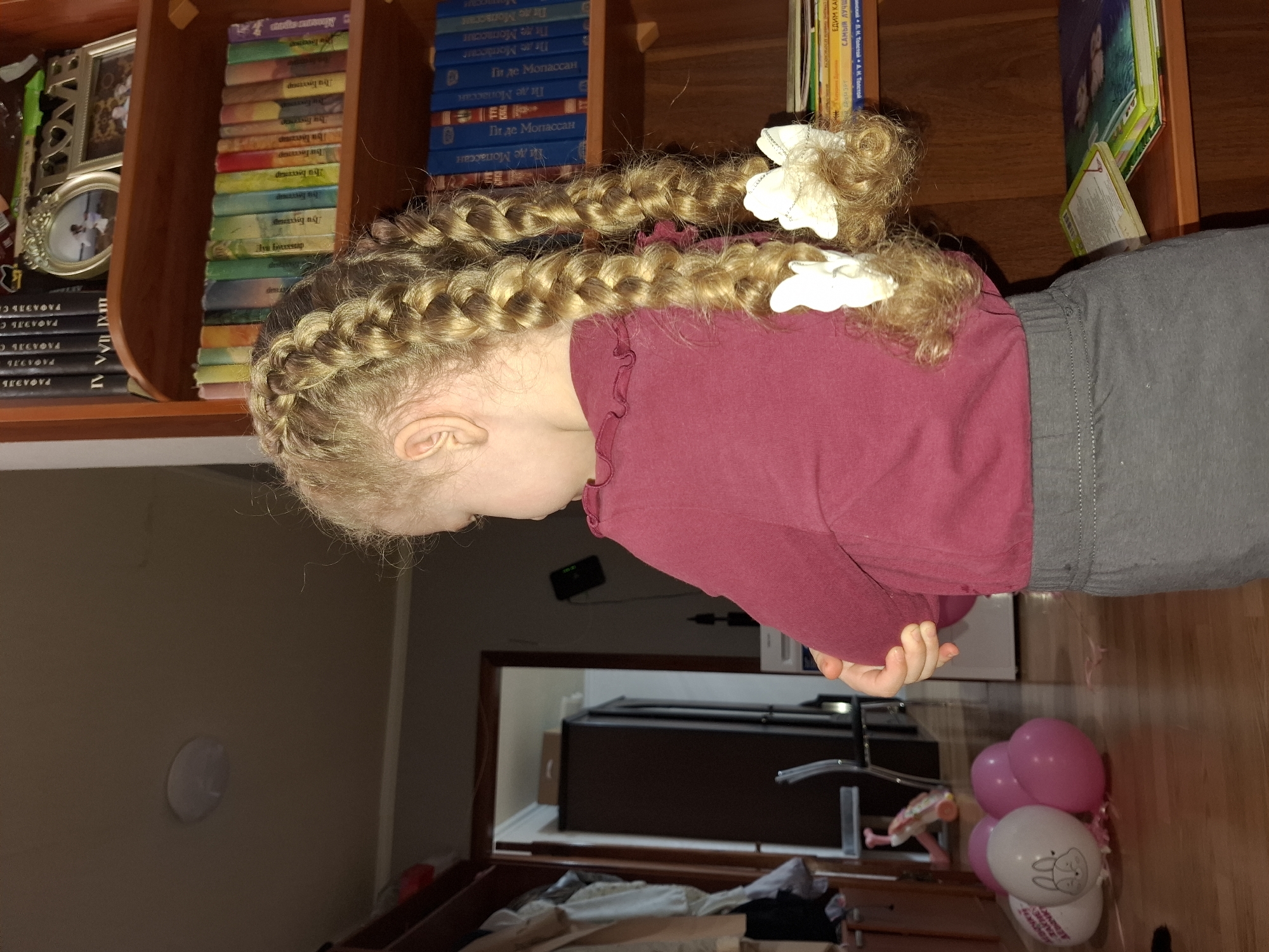 A young child, primarily seen from the back and side, is the central subject of this image, which is rotated 90 degrees counter-clockwise. The child has long, light blonde hair styled into multiple intricate braids, adorned with several white decorative bows. They are wearing a maroon long-sleeved top with subtle ruffle detailing on the shoulder seam and grey pants.
The setting appears to be indoors, likely a domestic environment in Chaikovsky, Russia, possibly a child's room or living area. To the left, a wooden bookshelf holds numerous books, many with Russian titles visible on their spines. Notable titles and authors include "ПИ ДЕ МОПАССАН" (Guy de Maupassant), "ЛУИ БУССЕНАР" (Louis Boussenard), "РАФАЭЛЬ" (Rafael), "В. Н. ТОЛСТОЙ. А. Н. ТОЛСТОЙ", and "САМЫЙ ЛУЧШ" (The Best). Other items on the shelf include decorative picture frames and a "HOME" sign.
In the lower right portion of the frame, a cluster of pink and white balloons rests on a light wooden floor. One white balloon prominently features a bunny illustration and the text "HAPPY BIRTHDAY". A mirror in the background reflects part of a dark piece of furniture and an open doorway, adding depth to the scene. The overall lighting is warm, suggesting an indoor setting with artificial light. The child's facial expression is not visible as they are looking away from the camera, seemingly downwards or towards something out of frame.