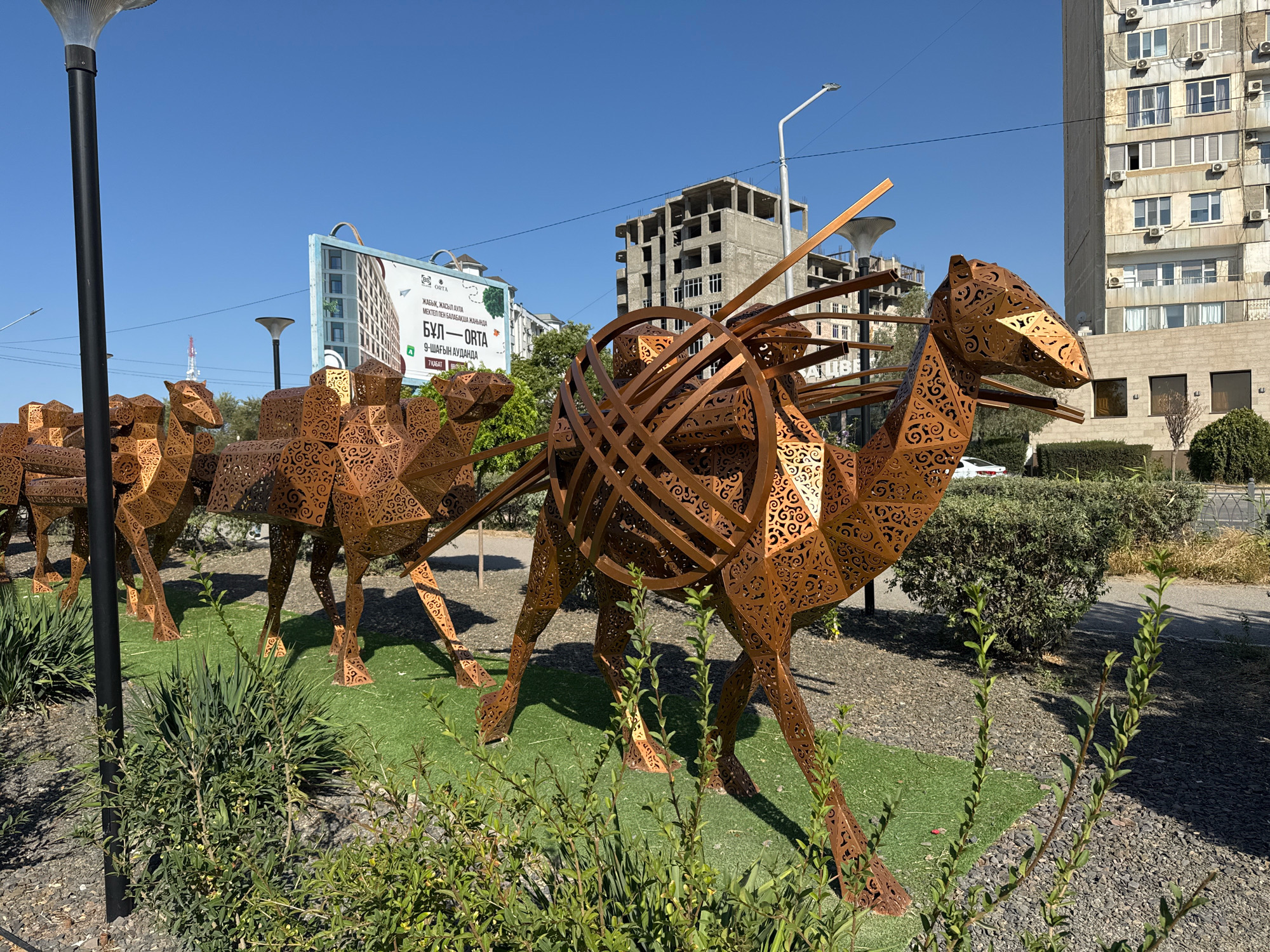 In Aktau, Kazakhstan, on a clear day, a series of large, ornate metal camel sculptures are displayed outdoors. The sculptures are made of a copper-colored metal with intricate, laser-cut patterns, giving them a delicate and detailed appearance. They are positioned in a row, with the lead camel significantly larger and more detailed than the others, featuring a unique, woven metal structure over its back, suggesting it is carrying a load or is a centerpiece. The sculptures are situated on a patch of artificial green turf, surrounded by gravel and low-lying green bushes.
In the background, a billboard advertises a residential complex named "ORTA," featuring an image of a modern building. Beyond that, unfinished and completed multi-story buildings, typical of urban development, are visible under a bright blue sky. A tall street lamp stands to the left of the frame, and other street lamps and a communication tower can be seen further in the distance. The scene suggests an urban environment with public art installations.
