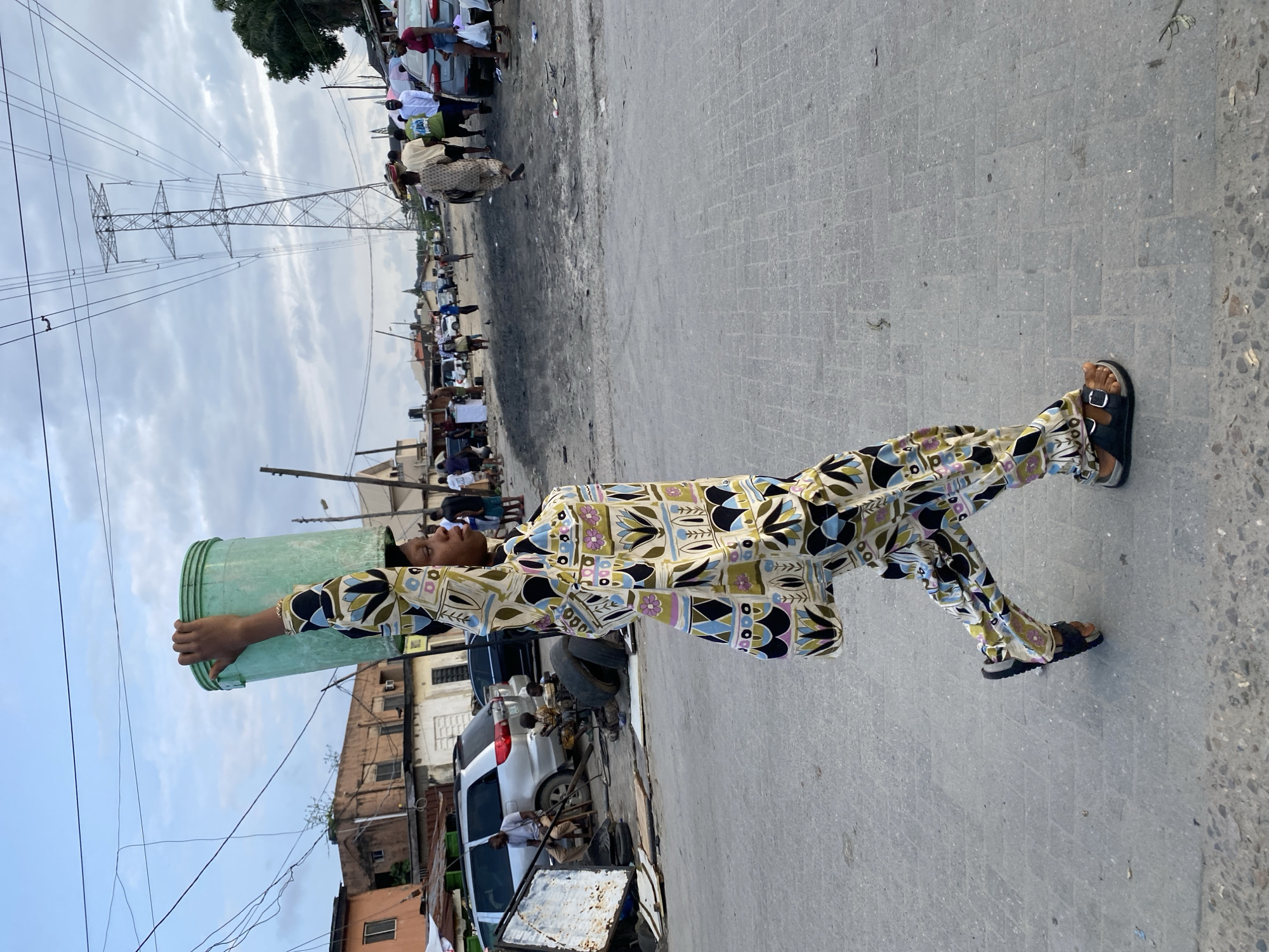 A young female, viewed in profile, walks across a paved road, balancing a large, light green plastic bucket on her head. She wears a vibrantly patterned, loose-fitting outfit composed of a long-sleeved top and matching trousers, both adorned with a repeating motif of abstract floral shapes in shades of cream, green, blue, black, and pink. She is also wearing dark-colored sandals. Her left arm is raised to steady the bucket, and her right arm hangs at her side, with a matching patterned bag slung over her shoulder.
The background depicts a busy, somewhat cluttered urban environment under an overcast sky. In the mid-ground, several cars are parked or driving on the road, including a silver SUV and a dark sedan. Scattered debris litters the road and the edges of the scene, suggesting a market or a working area. Buildings, some appearing to be made of concrete and others with corrugated metal roofing, line the street. Towering above the scene in the distance are large electrical pylons with multiple high-voltage wires stretching across the sky. Other people are visible walking and interacting in the background, some carrying items and others gathered in groups. The overall impression is one of daily life and activity in a bustling, possibly developing, urban setting.