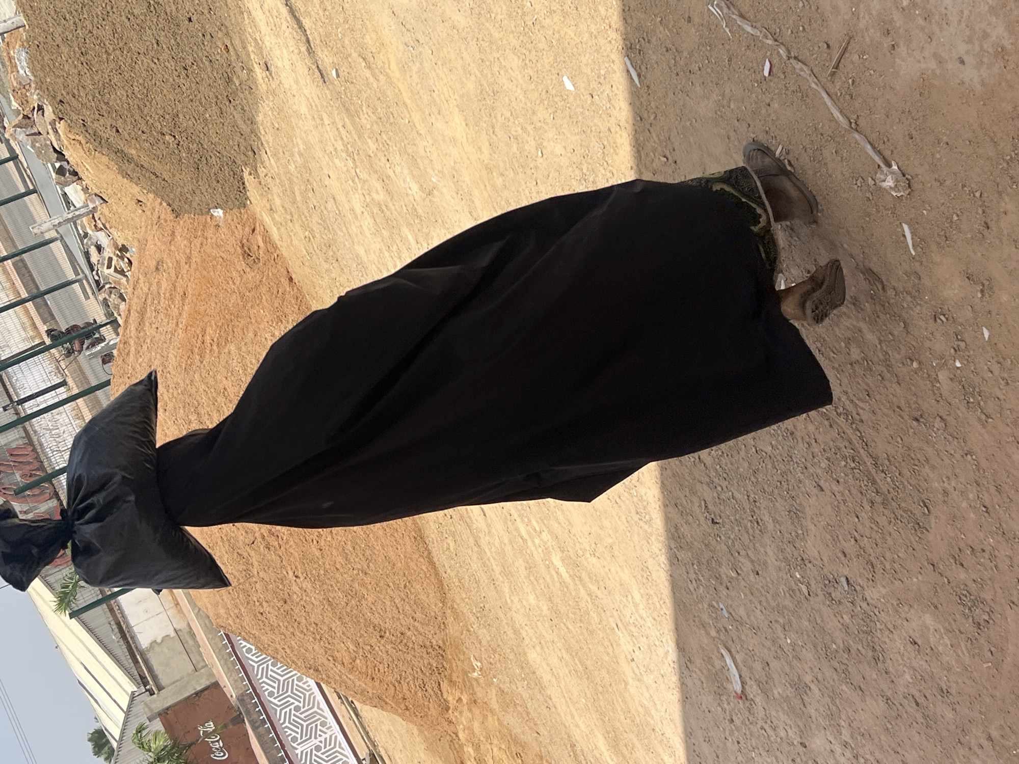 A person wearing a long black dress and carrying a black garbage bag over their head is walking on a dirt surface in Barakin Lerawi, Nigeria. The person is wearing sandals. A building and a fence are visible in the background.