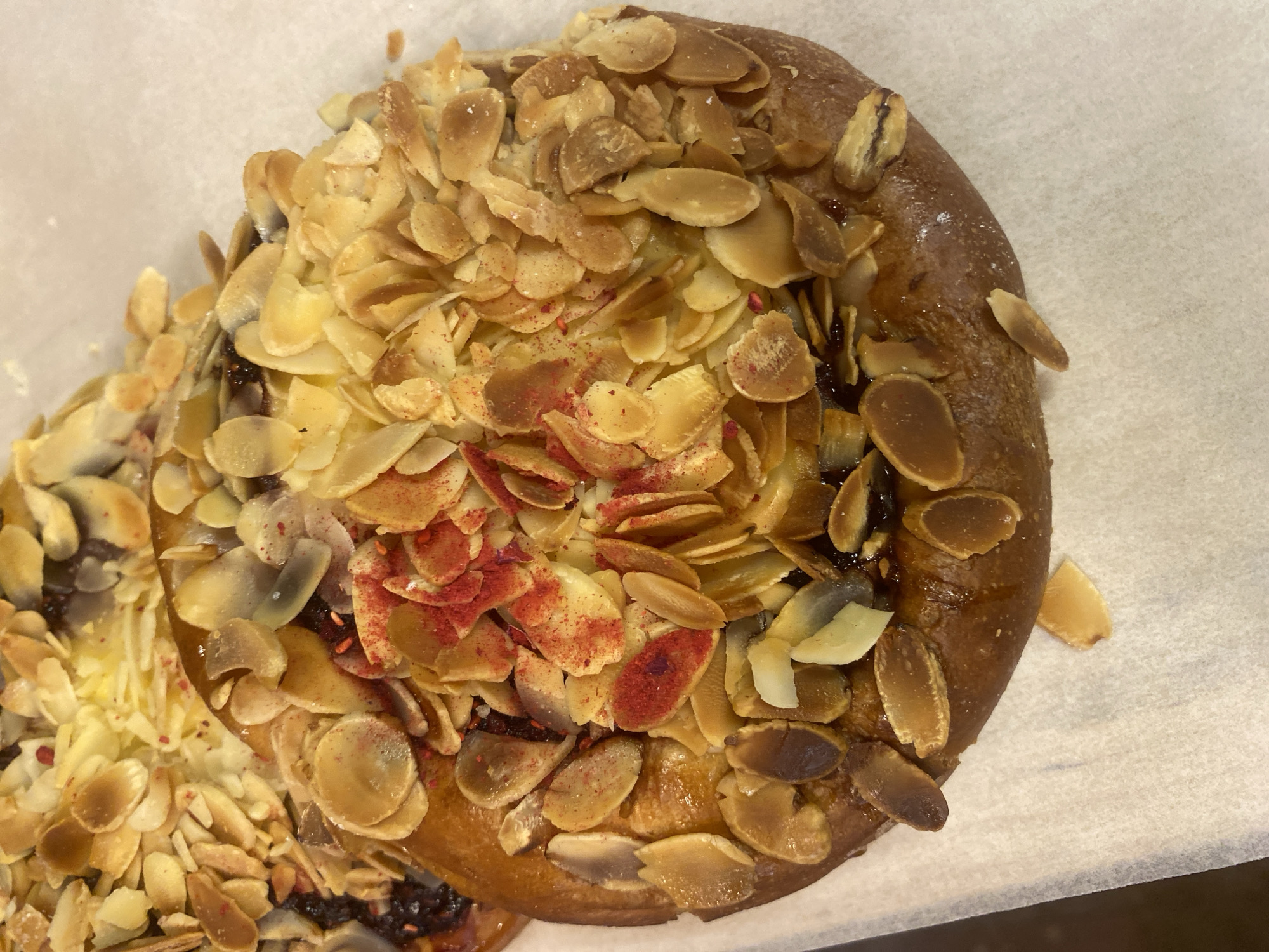 The image shows a close-up, overhead view of a pastry with sliced almonds and a red powder sprinkled on top. The pastry appears to be on a piece of white parchment paper, suggesting it is either being prepared or served. The lighting is somewhat dim, and there are no visible people or text. The pastry is golden brown with darker brown accents from the almonds. The red powder is visible in small clusters and appears to be a spice or topping. A portion of another, similar pastry is visible on the left side of the frame. The overall impression is of a baked good, possibly a dessert or breakfast item.