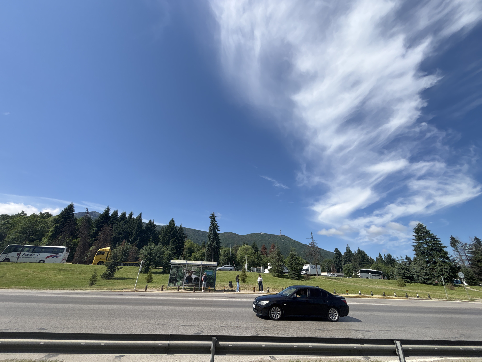 The image depicts a scenic view of a road and its surroundings in Sofia, Bulgaria. On the road, a black sedan is in motion, seen from its side profile, with a person visible inside the driver's seat. In the background, a grassy incline is covered with numerous coniferous trees, with some deciduous trees interspersed. A bus stop shelter with people standing around it is visible on the left side of the incline. Beyond the incline, a mountain range serves as a backdrop, with a clear blue sky overhead, featuring wispy white clouds. To the left of the incline, a white bus with "Fenix 2" written on its side is parked, and a yellow truck with a trailer is also visible. The overall scene suggests a sunny day in a semi-urban or suburban area with a natural landscape.