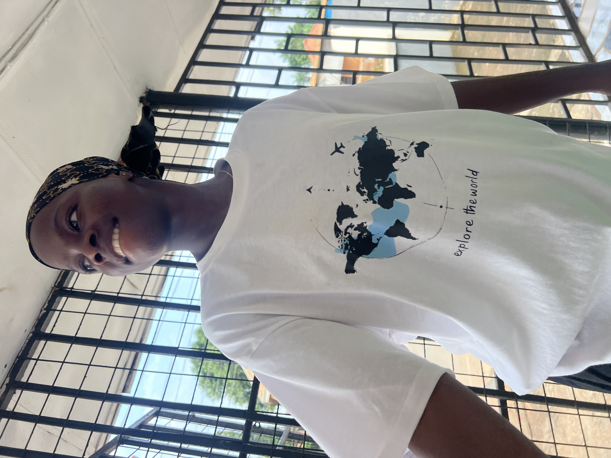 A young Black woman, wearing a white t-shirt with a world map graphic and the text "explore the world," stands in front of a barred window with a light-colored ceiling. She has a black and gold patterned headscarf on, and her gaze is directed upwards and to the right, with a bright smile. The background shows a glimpse of trees and buildings through the window, suggesting an outdoor or semi-outdoor setting in Nkpor, Nigeria. The lighting is natural and appears to be from an overhead source. The overall composition is a medium shot, focusing on the woman and her t-shirt.