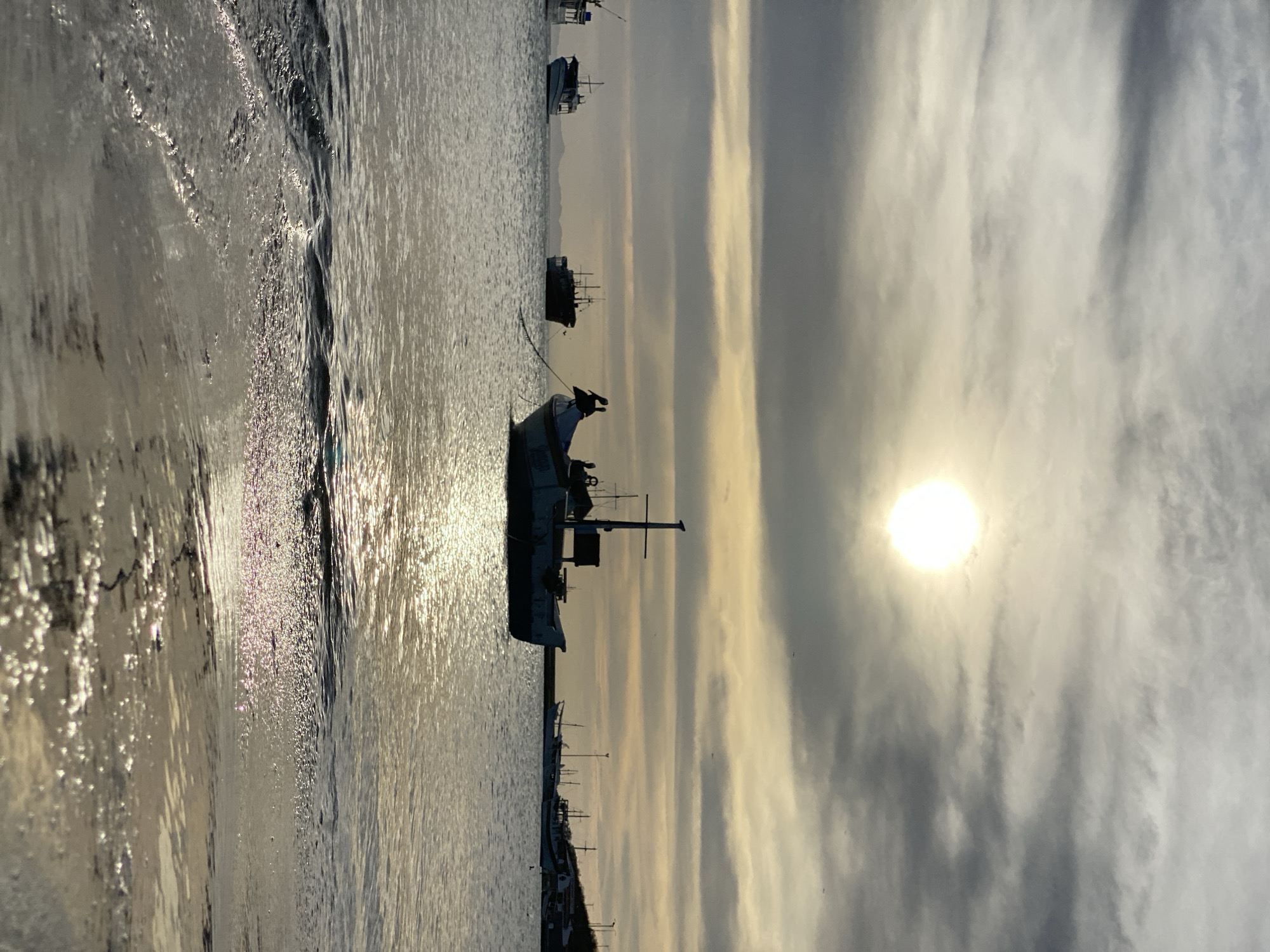 The image shows a body of water with several boats, likely a bay or harbor, with the sun shining through the clouds. The scene is located in the city of Juan Griego, Venezuela. The sun reflects off the water creating a glare and illuminating the waves. Several boats of various sizes are visible in the water, with at least one appearing to be moored or anchored. The sky is filled with clouds, obscuring much of the sky. Vegetation or structures are seen along the shoreline in the lower part of the image.
