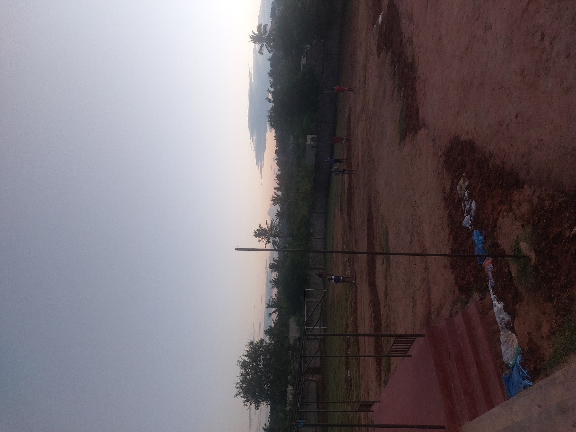 The image depicts a dusty field in Dar es Salaam, Tanzania, likely a recreation ground or sports field. In the background, palm trees and dense vegetation are visible against the twilight sky. Several people appear to be playing or walking on the field. A soccer goal and a netball pole stand on the field. A set of stairs on the right side of the image is strewn with garbage, including blue and white plastic bags. The lighting suggests it is either dusk or dawn.