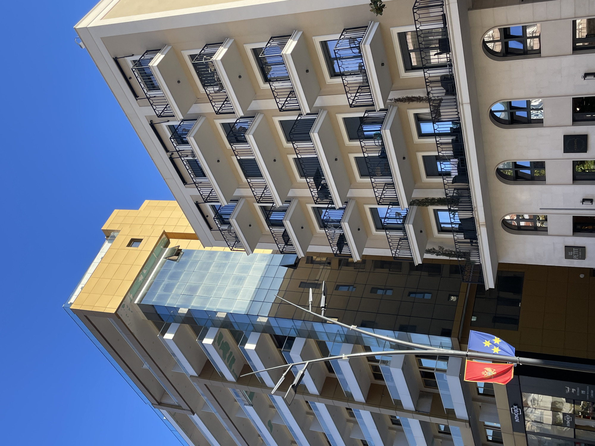 A low-angle perspective captures a modern building complex in Budva, Montenegro, under a clear blue sky. The buildings exhibit diverse architectural styles, including those with glass facades reflecting the sky and neighboring structures. The main building features multiple balconies with black wrought-iron railings. A flag of Montenegro and another flag, possibly of the European Union, hang from a street light in the foreground. Street-level retail establishments are suggested by signage and window displays in the lower portion of the image.