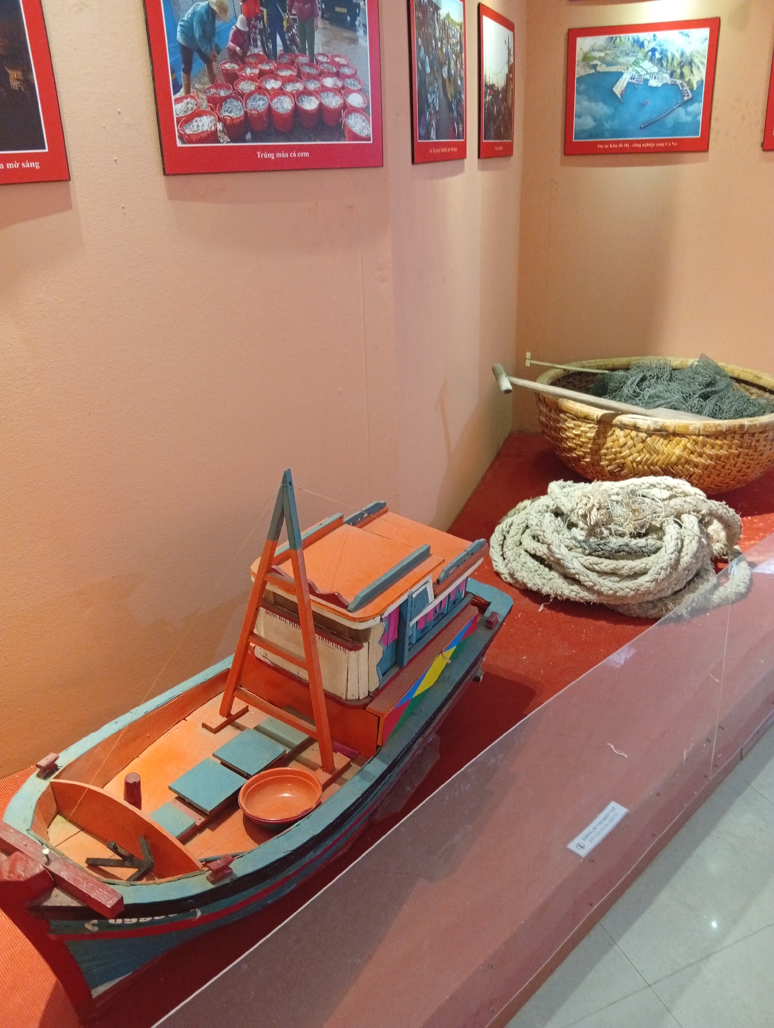 This image displays an indoor exhibit, likely within a museum or cultural center, focusing on maritime activities and regional development in Vietnam. In the foreground, a meticulously crafted model of a fishing boat, painted in vibrant shades of orange, blue, and green, rests on a red display surface. The boat features a small cabin, an anchor at the bow, and a red bowl on its deck. Adjacent to the boat, a large, coiled rope made of coarse natural fibers is presented, alongside a woven basket filled with a fishing net and a wooden oar or pole. A clear glass barrier runs along the front of the display, separating the exhibits from viewers.
The background wall, painted a light peach color, features several framed pictures. A prominent picture on the right shows an aerial view of a coastal development, identified by the Vietnamese text below it as "Dự án khu đô thị - công nghiệp cả Cà Ná" (Ca Na urban-industrial zone project). To its left, another framed picture depicts several people, presumably fisherfolk, attending to numerous red buckets brimming with small fish. The caption below this image reads "Trúng mùa cá cơm" (Good anchovy season), suggesting a prosperous anchovy harvest. Additional framed images are partially visible further to the left, showing more scenes of community or work.
Visible text on the model boat's cabin or an associated label includes "DONG BÀI, PHẦN" and "THÁP CHÀM", linking the exhibit to the Phan Rang – Tháp Chàm area. A label visible behind the glass barrier shows partial text like "SẢN XUẤT LỒNG BẪY HẢI" (Production of seafood traps) and "BAN QUẢN LÝ DỰ ÁN" (Project Management Board). The display effectively combines elements of traditional fishing practices with representations of modern industrial development plans for the coastal region. The indoor setting provides no specific cues for the time of day or weather, and the exhibited objects do not convey emotions.