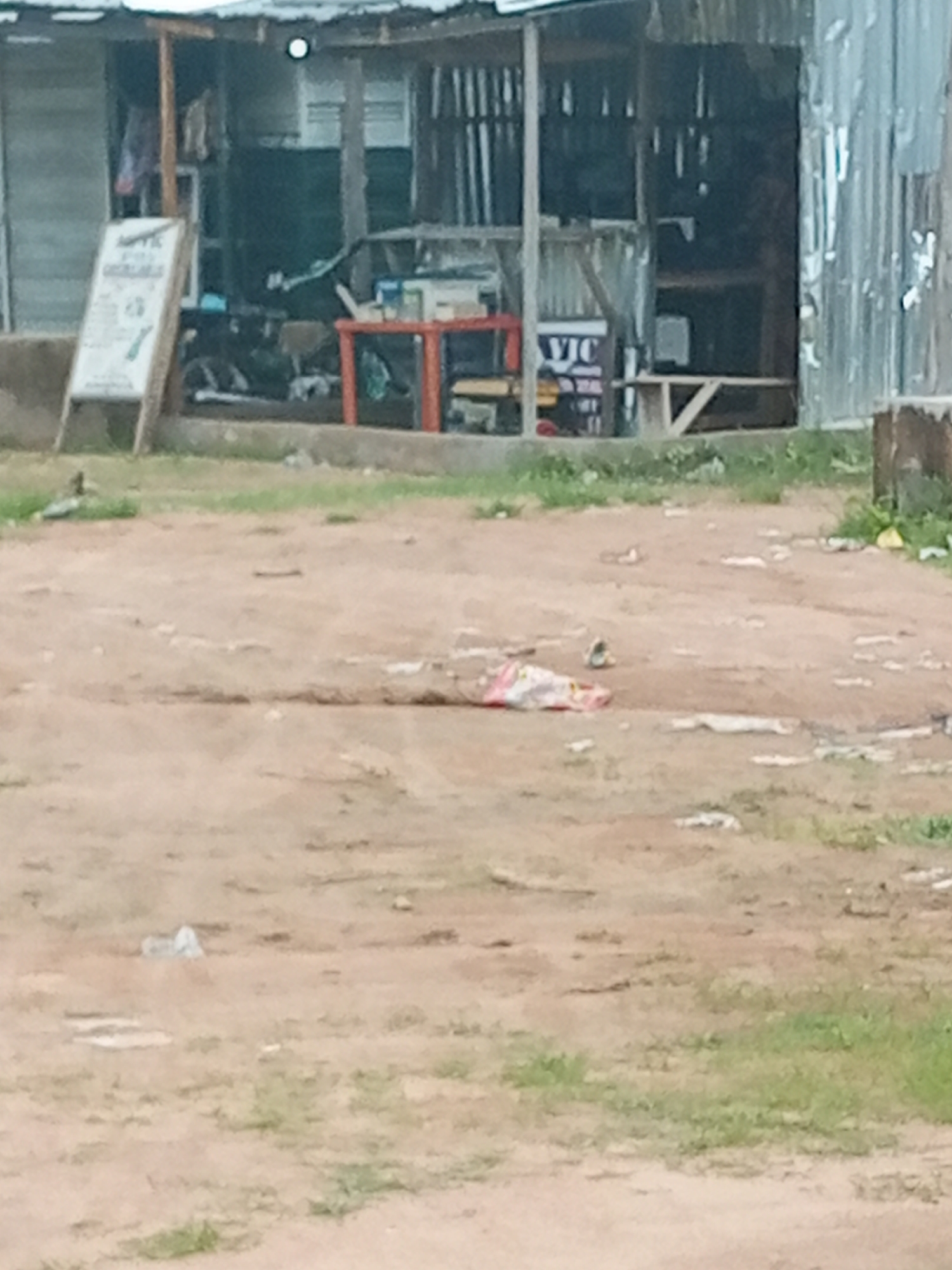 The image depicts an outdoor scene in Jalingo, Nigeria. In the foreground, a dirt and gravel area is scattered with small pieces of debris and some patches of dry grass. A white plastic bag and other small litter are visible on the ground.
In the background, there is a makeshift structure resembling a shop or market stall. It appears to be constructed from corrugated metal and wood. The stall has an open front, revealing various items inside, though they are not clearly discernible due to the image quality. To the left of the stall, a leaning sign is propped up. The sign is white with dark text and graphics, including what appears to be a recycling symbol. Partially visible text on the sign includes "VIC".
Within the stall area, a red table or counter is visible. Behind the table, there are some objects that could be appliances or goods for sale, as well as shelving. A bicycle or motorcycle is parked near the stall entrance. The overall setting suggests a commercial area in a town or city, likely a marketplace or a roadside vendor. The lighting suggests it could be daytime, but no specific time can be determined. The weather appears to be dry.