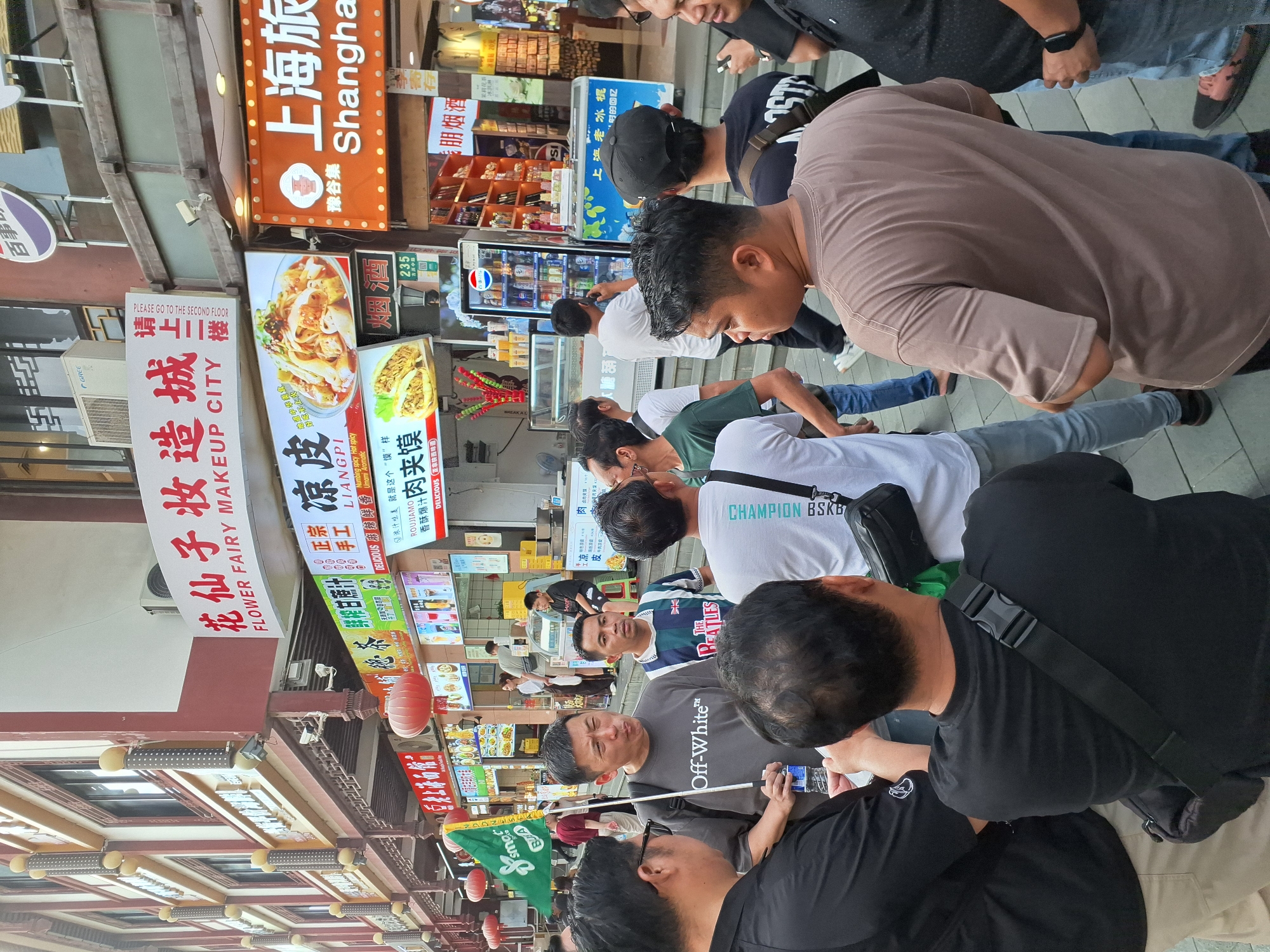 In an outdoor pedestrian area in Shanghai, China, a group of men are gathered. The scene is set against a backdrop of shops with illuminated signs. One sign reads "FLOWER FAIRY MAKEUP CITY" and another displays "Shanghai." In the foreground, several men are conversing. One man, wearing a dark grey "Off-White" t-shirt, holds a green flag and a bottle of water. Another man wears a t-shirt with "The Beatles" and a Union Jack emblem. Behind them, other individuals are visible, some looking at their phones. The ground is paved with grey tiles. A row of shops features food stalls, a Pepsi vending machine, and shelves stocked with goods. Red lanterns hang from the buildings. The atmosphere appears to be casual and social.