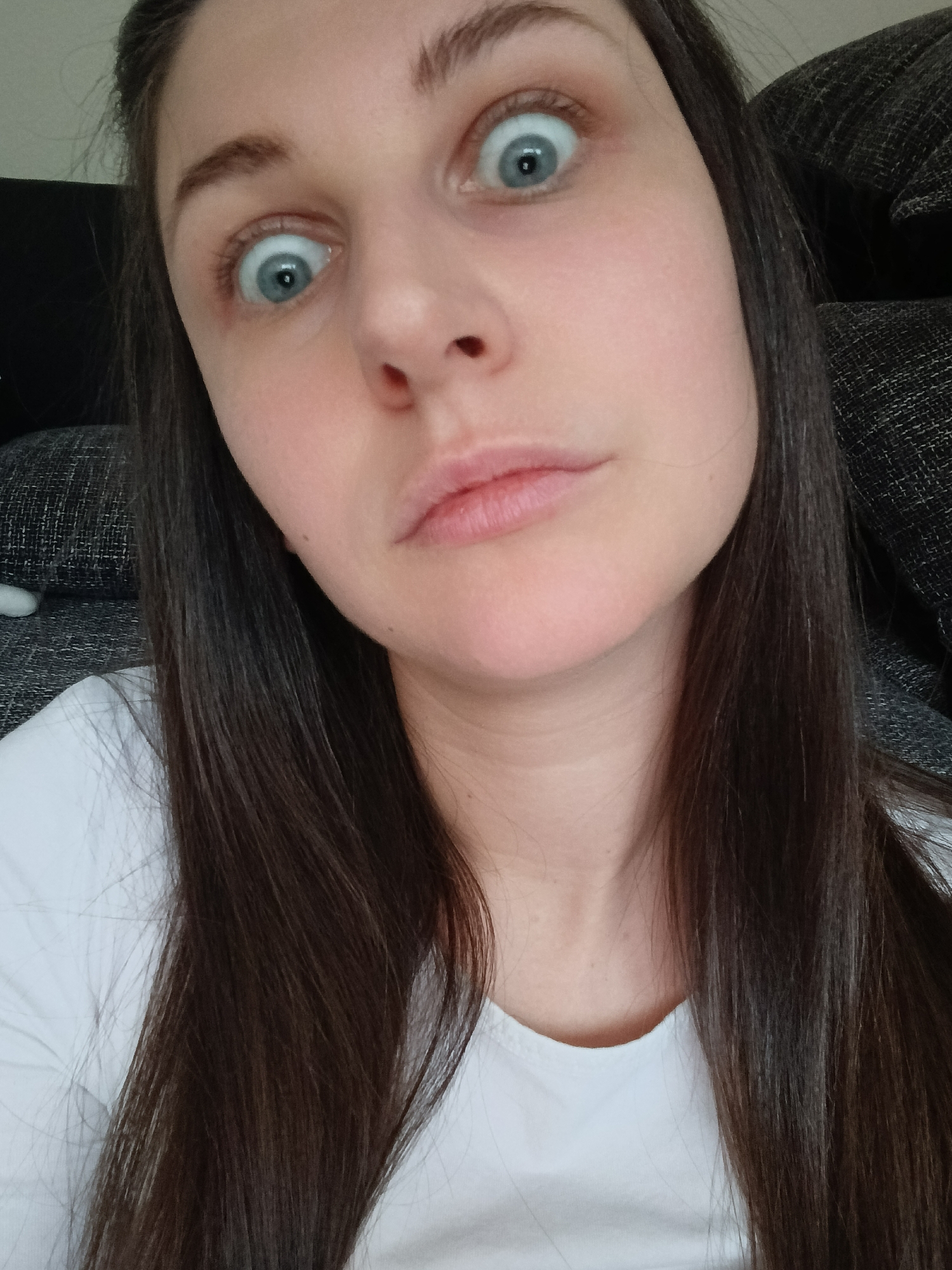 A close-up selfie depicts a woman with fair skin, long dark brown hair, and light blue eyes. Her eyes are wide and her eyebrows are slightly furrowed. Her lips are closed with a slight pout. She is wearing a white shirt. The background is out of focus, revealing a dark grey textured surface. The city and specific location are unknown.