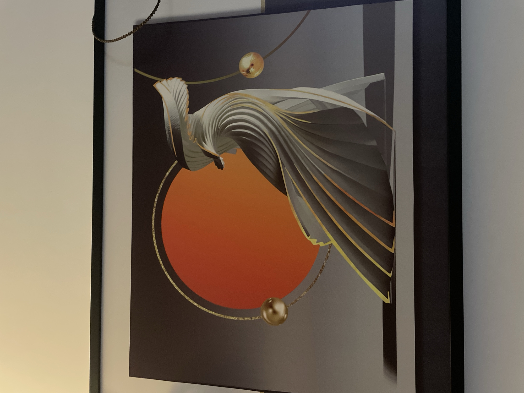 The image is a close-up shot of an abstract art piece, likely a print or painting, framed in black. The artwork features a gradient background transitioning from a dark grey to a lighter grey. A large orange circle dominates the center of the composition. Overlaid on the circle is a swirling, sculptural form that appears to be made of layered segments, possibly representing feathers or wings. This form has both light and dark grey tones, accented with gold lines along the edges of the segments. Gold spheres are positioned along thin, curved lines within the artwork.
The location of Bac Nha Trang Ward, Vietnam, is not depicted in this image. No people, specific objects, actions, interactions, or further environmental details are visible other than the wall on which the artwork hangs.