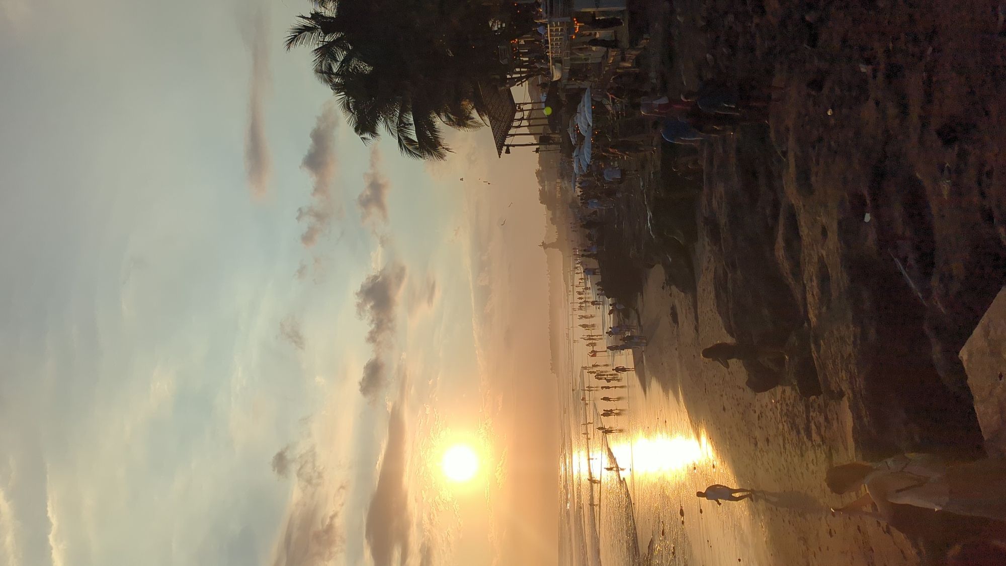 This media file captures a vibrant sunset scene on a popular beach in North Kuta, Indonesia. The image, originally rotated, shows a broad stretch of sandy shore meeting the ocean under a colourful sky.
**Subjects:** The central subject is the setting sun, a brilliant golden orb low on the horizon, casting a powerful warm glow. Numerous people are visible along the beach, mostly in silhouette, walking along the waterline or standing in small groups. Their elongated reflections are prominent on the wet, reflective sand. To the left, dark silhouettes of palm trees frame the upper portion of the scene. Further back from the immediate shoreline, a bustling area with various beachside establishments, including structures with roofs, umbrellas, and seating, can be discerned. To the right, the beach rises into a darker, possibly rocky or sandy, bank. The sky above is painted with soft blues, purples, and oranges, with scattered clouds catching the last light of the day. A few small birds are visible in the distance.
**Activity or event taking place:** The scene depicts an evening gathering where people are enjoying the sunset at a tropical beach. Many individuals are leisurely strolling along the water's edge, taking in the picturesque view, while others appear to be relaxing or dining at the beachside establishments. The atmosphere suggests a popular recreational spot.
**Notable details:** The time of day is clearly sunset, characterized by the low sun, the dramatic warm colours dominating the sky and reflections on the water and sand. The weather appears clear and pleasant, with gentle clouds and calm waters. The presence of palm trees, beach cafes, and the general ambience are strong location cues for a tropical beach destination like North Kuta, Indonesia. The overall emotion conveyed is one of relaxation, enjoyment, and appreciation for natural beauty. No discernible text is visible in the image.