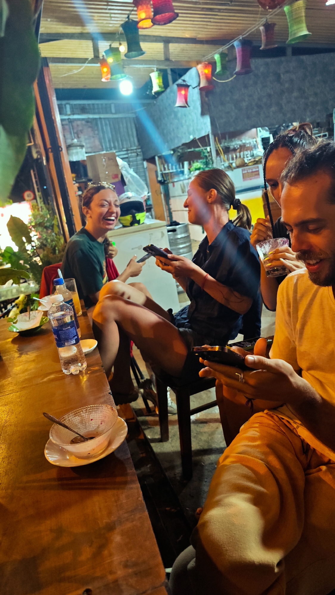 This image depicts a lively evening scene at a casual outdoor or semi-outdoor establishment, likely in Mù Cang Chải, Vietnam. Four individuals—three women and one man—are seated around a rustic wooden table, engaged in conversation and with their phones.
In the foreground, a man with a beard, wearing a light yellow t-shirt, smiles as he looks down at his smartphone. Beside him, a woman in a dark shirt is also focused on her phone, her gaze directed to the left. Another woman, partially obscured behind the man, holds a drink with a straw. To their left, a woman in a dark green t-shirt laughs heartily, her head tilted back, looking towards the woman next to her. The group's expressions convey joy, relaxation, and friendly interaction.
On the wooden table, a clear plastic bottle of "Aquafina" water, a glass with an orange liquid, and an empty bowl with a spoon are visible. The background reveals a charming and informal setting, adorned with strings of colorful, decorative lanterns hanging overhead, providing warm illumination alongside a bright, unshaded bulb. Elements of a bar or kitchen area can be seen, including metal beer kegs, a cooler or refrigerator, and a white and yellow motorcycle helmet. Green foliage lines the left side of the frame, contributing to the outdoor ambiance. The time of day is clearly evening or night, given the artificial lighting and dimness.