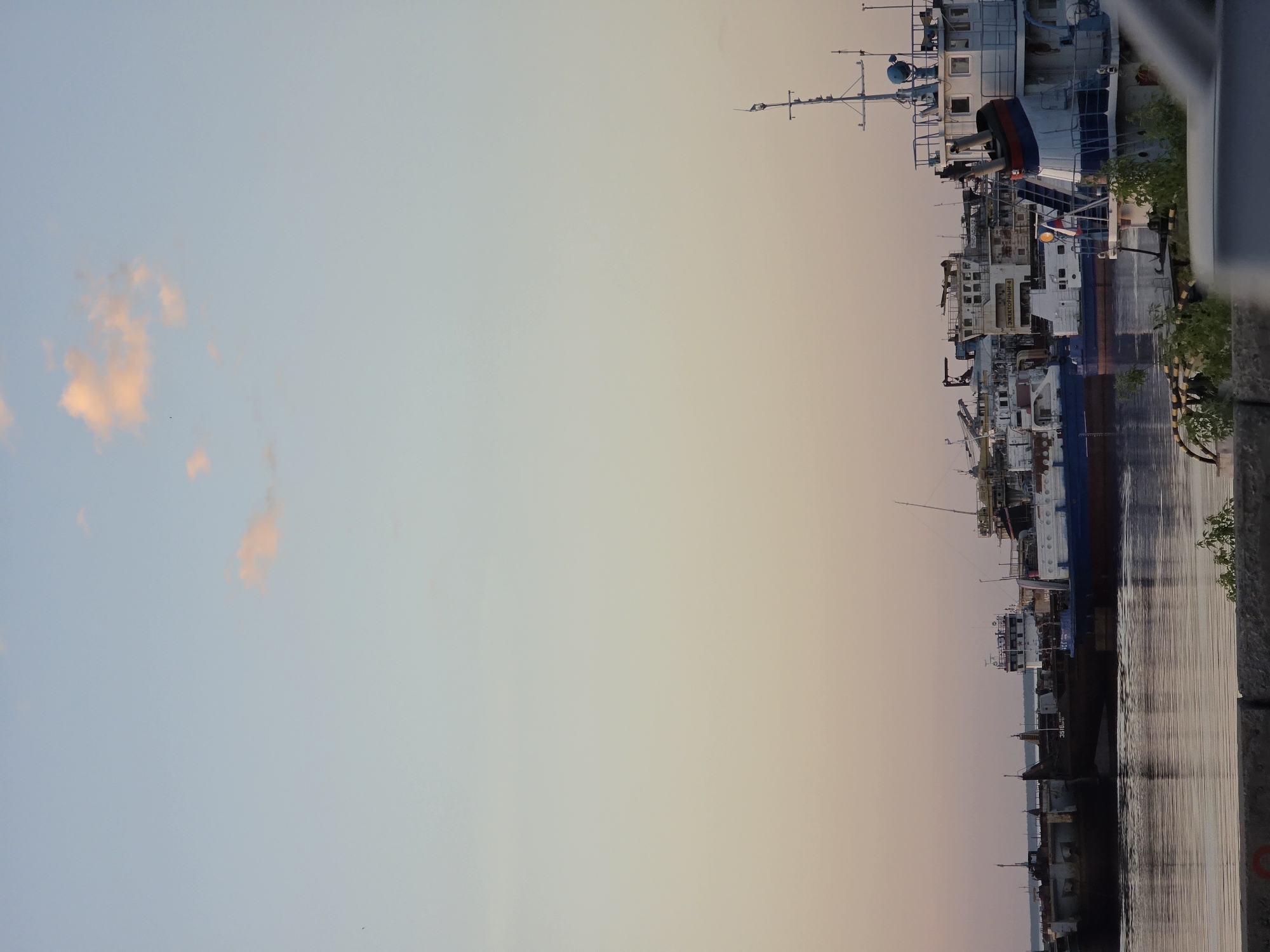The image shows several ships docked along a waterfront at sunset. The location is Chaikovsky, Russia. The sky is a gradient of blue and orange. Several ships are docked parallel to each other, with detailed superstructure visible. Reflections of the ships are visible in the water.