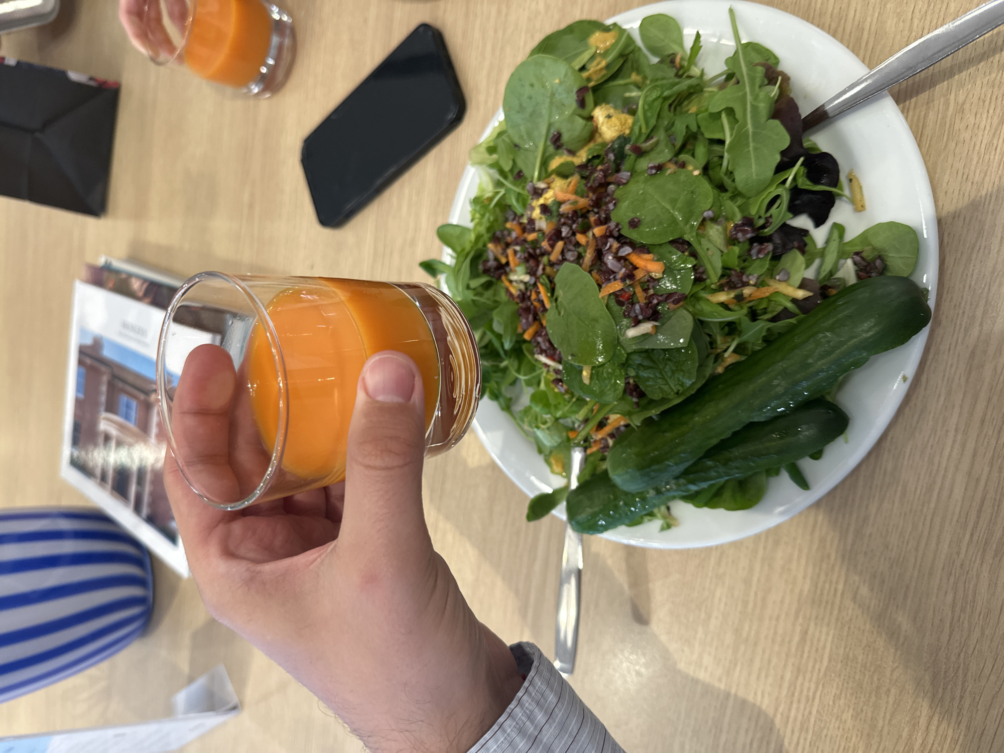 A view from above captures a person holding a glass of orange juice next to a white plate of salad with cucumbers. The scene appears to be in an indoor setting with a wooden table surface. A smartphone and another glass of orange juice are visible in the periphery, along with printed materials and a decorative striped vase. The person's arm is visible, clad in a light-colored shirt.