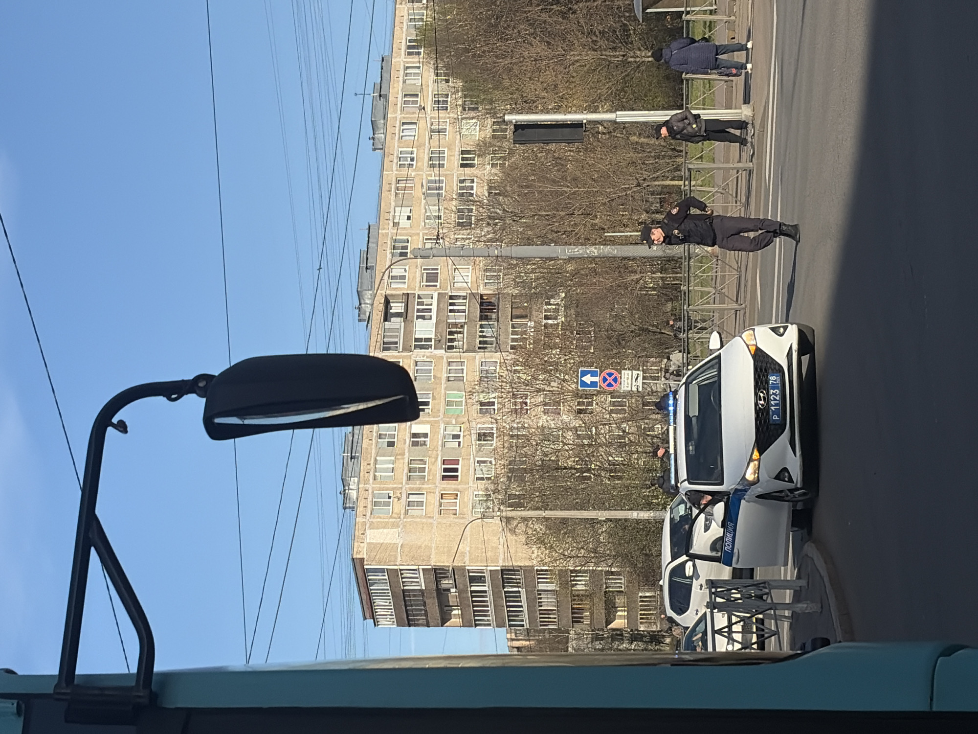 This image, taken from inside a vehicle on a bright, clear day, captures a street scene in Saint Petersburg, Russia. The perspective is slightly elevated, looking out onto a road and a multi-story apartment building in the background. The sky is a vivid blue, and overhead electrical wires crisscross the frame.
The central focus is a white Hyundai police car with its front passenger door open, parked on the side of the road. The word "ПОЛИЦИЯ" (POLICE) is visible on its side door, and its Russian license plate, "P 1123 78," confirms the Saint Petersburg location. Blue flashing lights are visible on the car's roof.
Several individuals are present. One police officer in a dark uniform and cap stands in the foreground, near the police car, seemingly engaged in an observation or interaction. Another officer is visible further down the street, standing near a traffic light pole. At least one more officer is partially visible near the police car. Behind a metal pedestrian barrier, two other individuals, possibly civilians, are standing, one in a dark blue jacket and the other in a black puffer jacket.
The setting includes a typical Soviet-era apartment building with numerous windows, bare trees suggesting early spring or late autumn, and various street furniture elements like metal barriers and a street sign. The sign depicts a blue square with a white left-pointing arrow and a red circular sign, possibly indicating traffic rules. The presence of police officers and a police vehicle suggests either a routine patrol, a traffic stop, or a response to a specific event. No overt signs of distress or conflict are visible; the scene appears calm under the sunny sky.