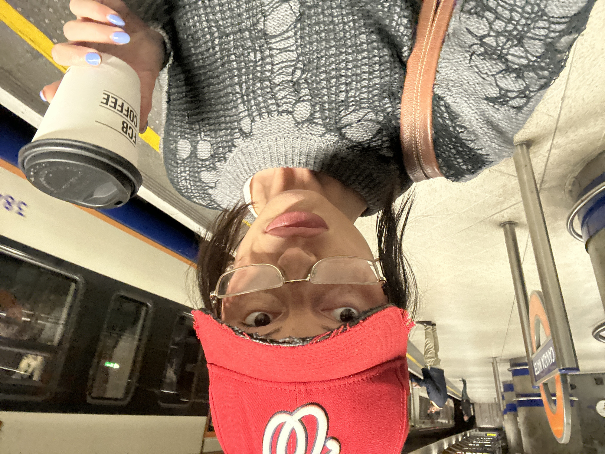 A person is taking a selfie at a London Underground station. The station has a sign indicating it is Camden Town, a station on the Northern Line of the London Underground. A train is visible on the left, and other people are further down the platform. The person is wearing glasses, a red baseball cap with a logo, a gray sweater, and has pale blue nail polish. They are holding a coffee cup from FCB Coffee. The image is upside down.