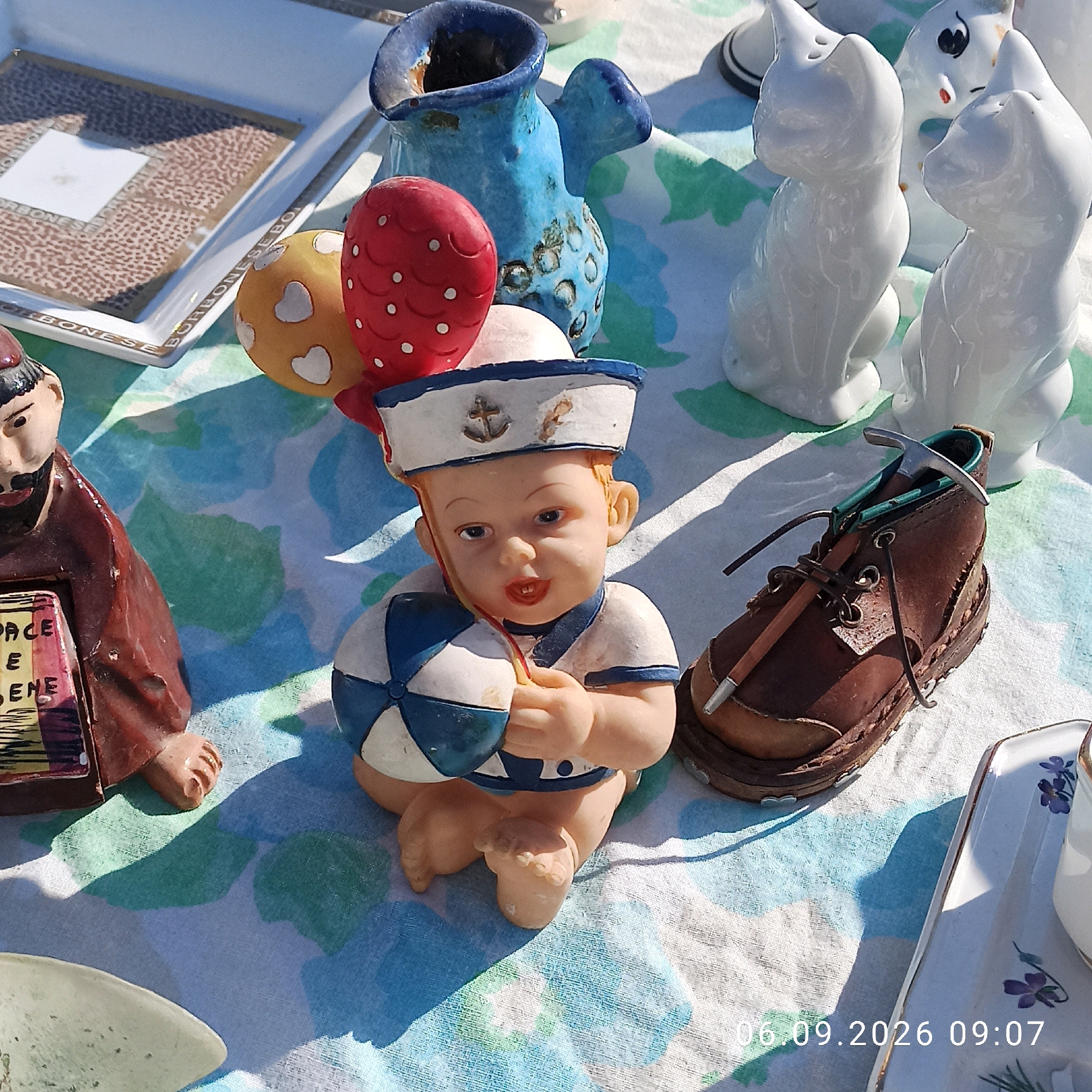 The image displays an outdoor scene, likely a market or yard sale, set upon a patterned tablecloth with blue and green abstract shapes. In the foreground, a ceramic figurine of a baby in a sailor outfit holds a blue and white striped ball. To the left of the figurine is another ceramic figure, appearing to be a monk or friar, holding a red plaque with white text. Behind these figures are various other ceramic objects, including a blue pitcher with textured detailing, two white ceramic cat figurines, and a small brown leather shoe with a metal pickaxe-like object inserted into it. Further to the right, a white ceramic tray with a floral design is partially visible. The background shows a flat surface with some texture. Sunlight casts shadows across the scene. The timestamp "06.09.2026 09:07" is superimposed on the lower right portion of the image, suggesting a date and time. The overall impression is of a collection of decorative items arranged for display. The location of Sannicandro di Bari, Italy, is not visually identifiable from the image content alone.