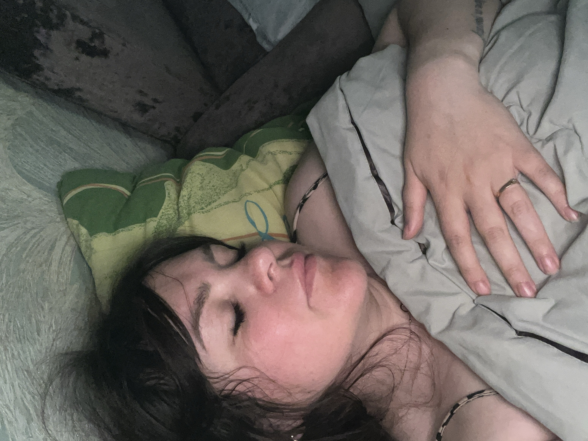 A woman is lying down, presumably sleeping, on a surface covered with bedding. She has dark hair and is partially covered by a light-colored blanket. Her hand, adorned with a ring, rests on the blanket. The background appears to be a couch or other upholstered furniture, in a dimly lit setting. There is no indication of this being in Noviy, Russia.