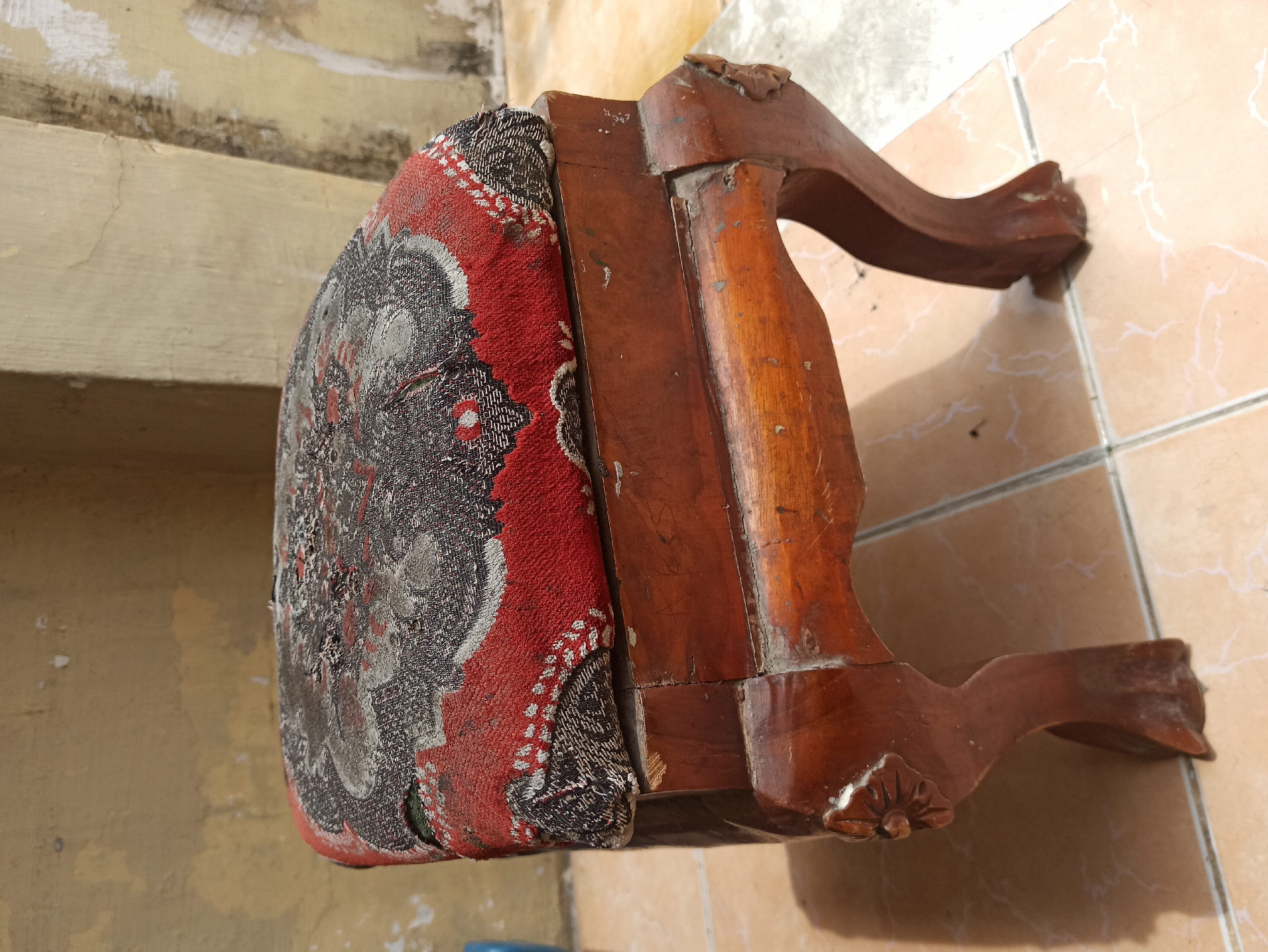 A close-up, eye-level shot shows a small, wooden stool with a patterned fabric cushion. The stool has four curved legs, each with a carved floral design on the upper portion. The wood is a dark brown, with visible wear and tear, including scratches and peeling varnish. The cushion is upholstered in a fabric with a red base color and a complex pattern of grey, black, and white. The fabric appears worn and slightly torn in places. The stool is placed on a tiled floor, which is light brown with some white veining. In the background, a textured, light yellow wall is visible, with patches of peeling paint. The scene suggests an outdoor setting, possibly a courtyard or a patio, given the tiled floor and the wall's appearance. The lighting indicates it is daytime, with shadows cast by the stool on the floor. The image does not contain any people. The location is identified as the City of Medan, Indonesia.