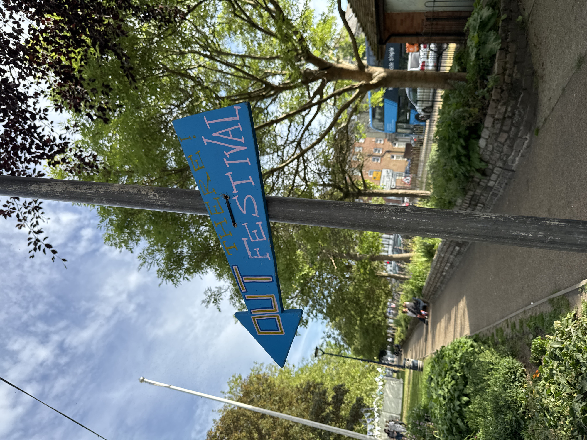 Here's a description of the image:
The image shows a close-up of a blue, arrow-shaped sign attached to a gray metal pole. The sign reads "OUT THERE FESTIVAL!" in pink and yellow lettering, and the arrow points downward. The pole is positioned in an outdoor setting, likely Great Yarmouth, United Kingdom. In the background, there's a tree with green leaves, a building, and a glimpse of a blue double-decker bus. The scene is sunlit with some cloudy skies visible. A path and greenery are visible in the background.