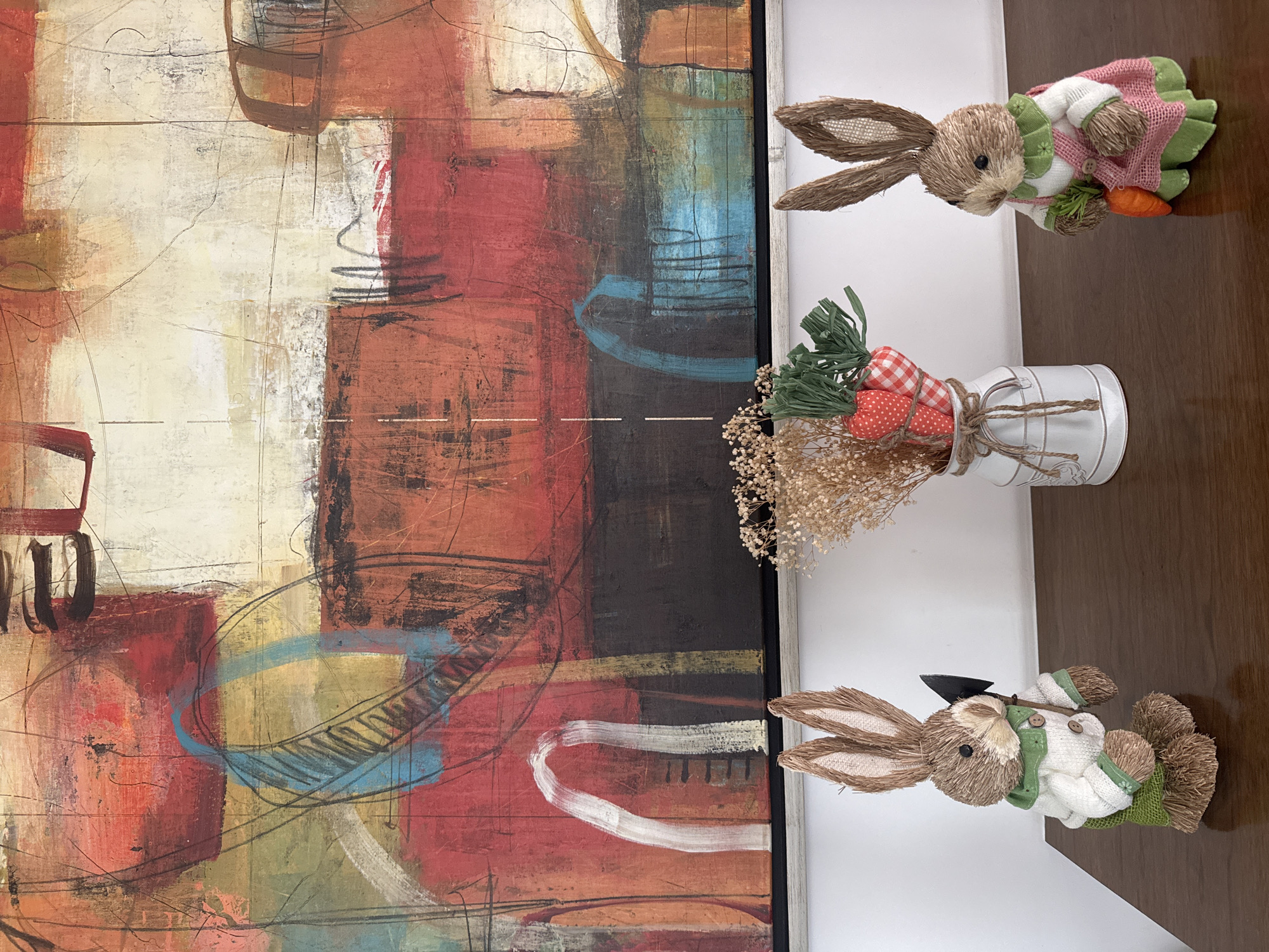 The image shows a section of wall, and a tabletop display. The wall features a large abstract painting with a mix of colors, including red, brown, black, white, and yellow. The painting appears to be framed in a light-colored frame. The tabletop is a brown, smooth surface. On the tabletop, there are two decorative straw bunny figurines, one with a pink dress and one with a gardening shovel, and a small metal container filled with carrots and artificial foliage. The setting appears to be indoors. The location information provided (San Bernardino Tlaxcalancingo, Mexico) cannot be verified within the image.
