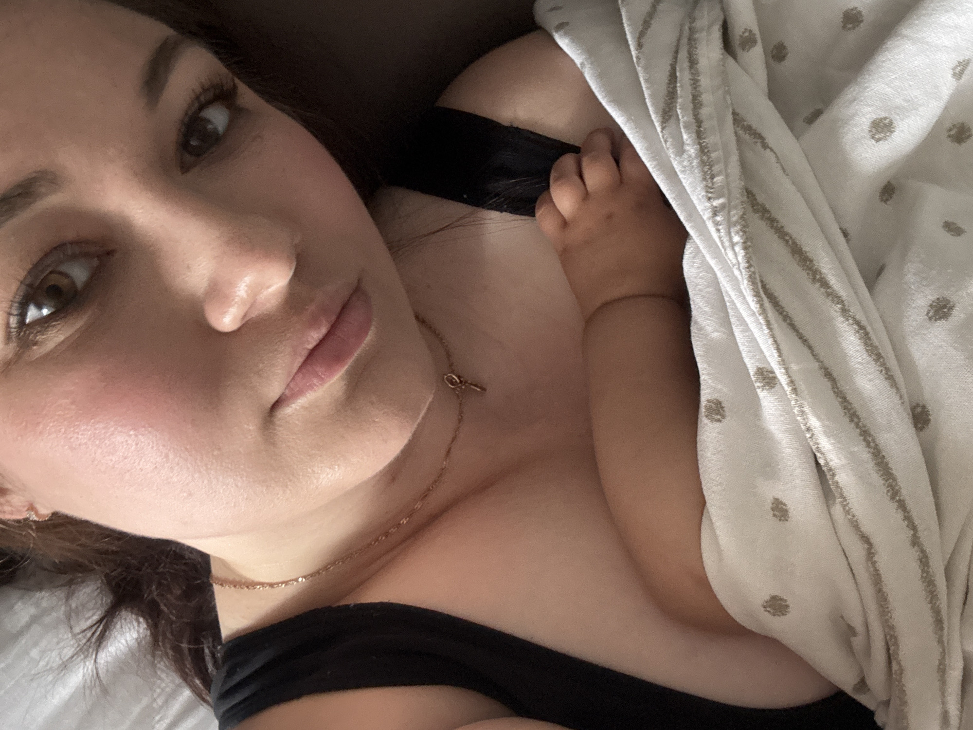 This is a close-up, first-person perspective selfie of a young woman lying down. She is wearing a black tank top and a thin gold necklace with a small key pendant. Her head is tilted, and she is looking slightly off-camera. A baby's hand is visible resting on her chest, partially covered by a white and beige striped blanket with small polka dots. The background is dark and indistinct, suggesting an indoor setting, likely a bedroom. There is no information in the image to indicate a specific location in the city of Chaikovsky, Russia.