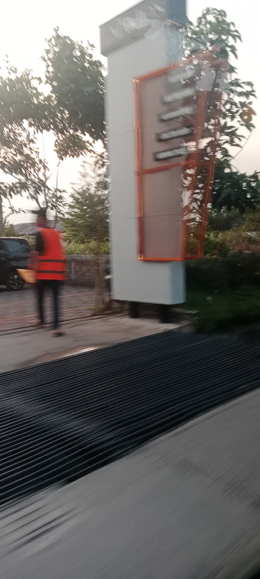 In Banyuraden, Indonesia, an outdoor scene captures a person wearing an orange vest and dark pants. They are standing near a black car and a tall white pillar that has an orange, mesh-like structure with lights on it. In the foreground, a dark, ridged surface is visible, possibly a drain grate or a textured path. Trees and foliage form the background, with a soft sky overhead, suggesting it might be early morning or late afternoon.